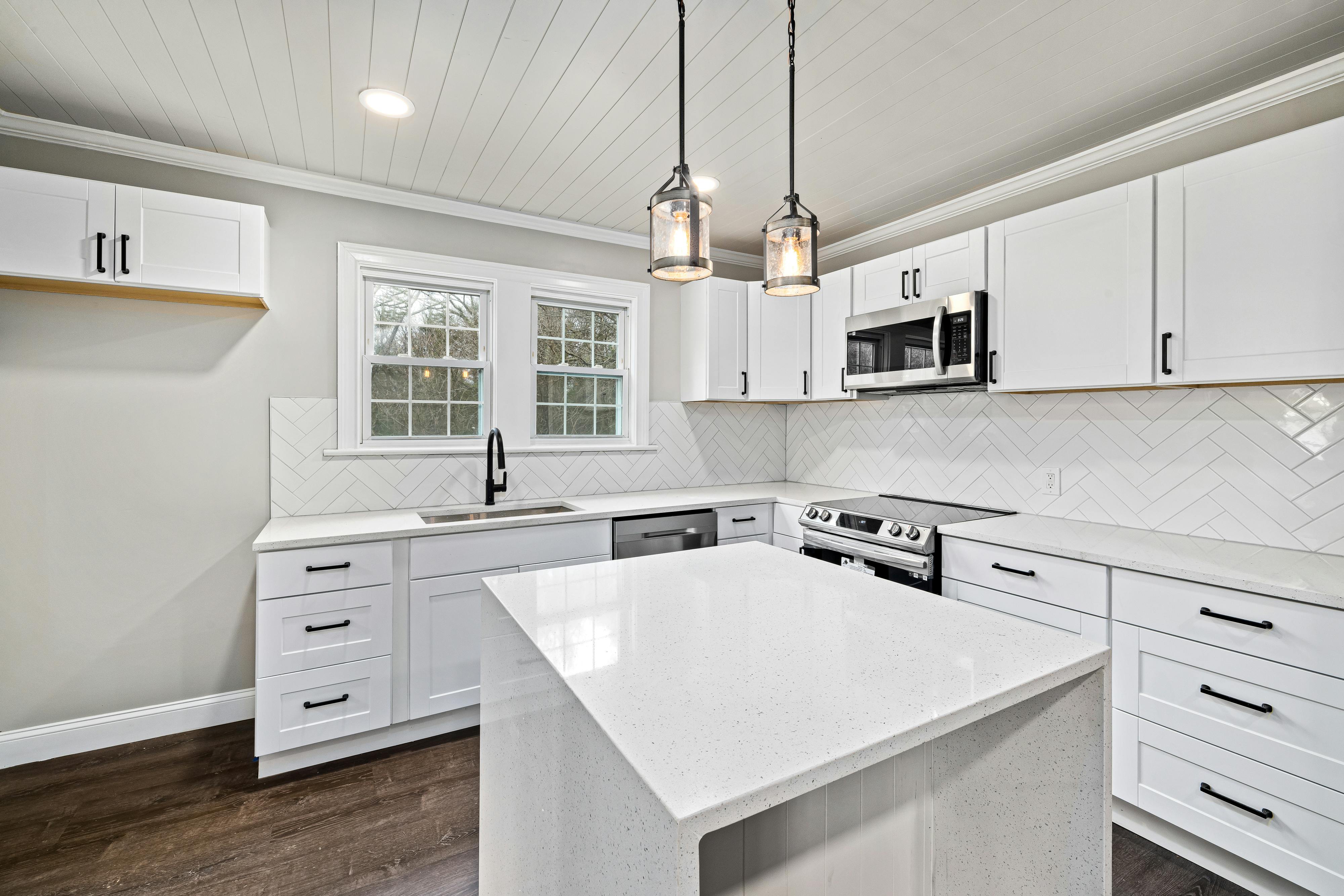 Modern white kitchen with an island, stainless steel appliances, black fixtures, and herringbone tile backsplash.