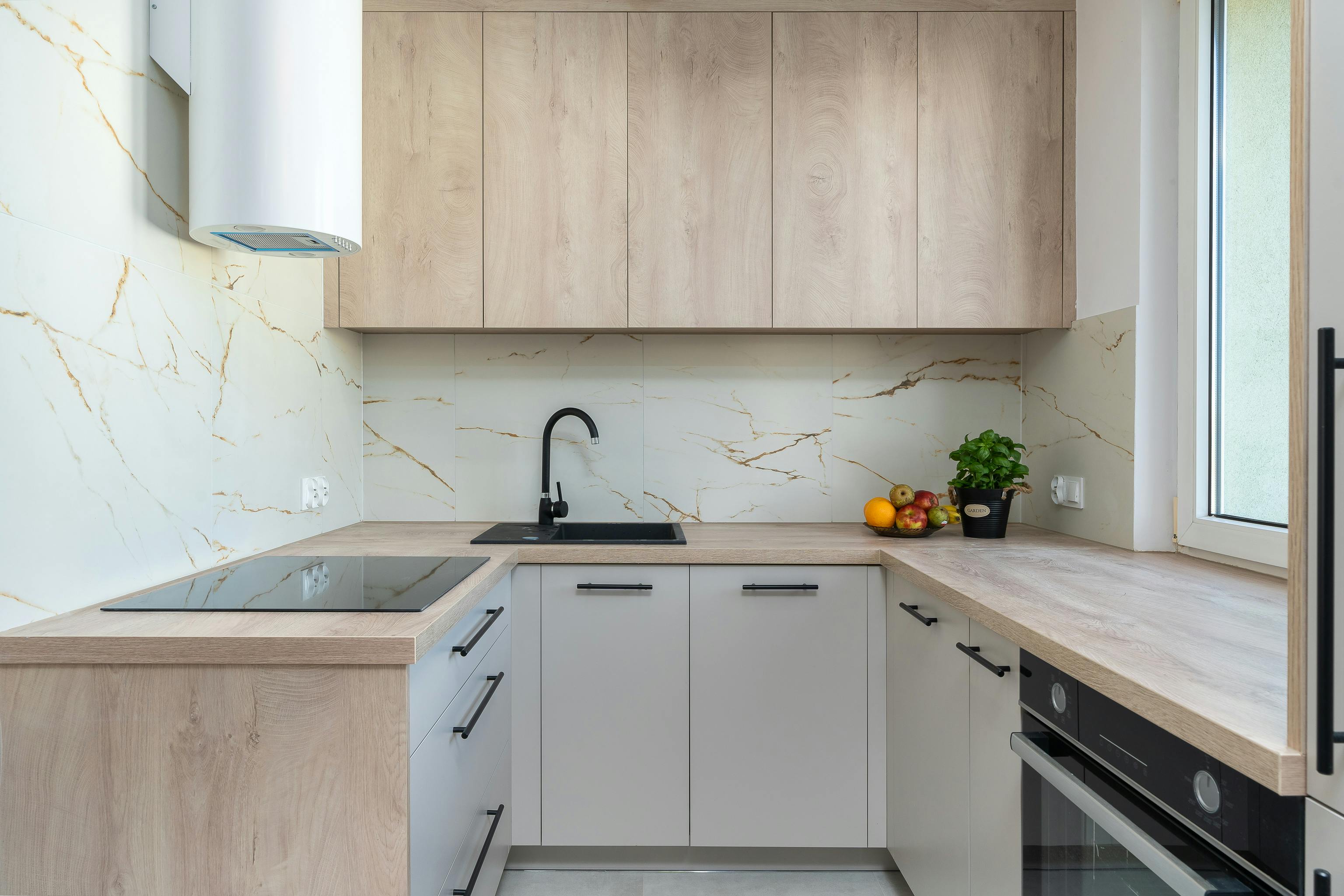 Modern kitchen with light wood cabinets, marble-patterned backsplash, black faucet, induction cooktop, and a bowl of fruit next to a potted plant.