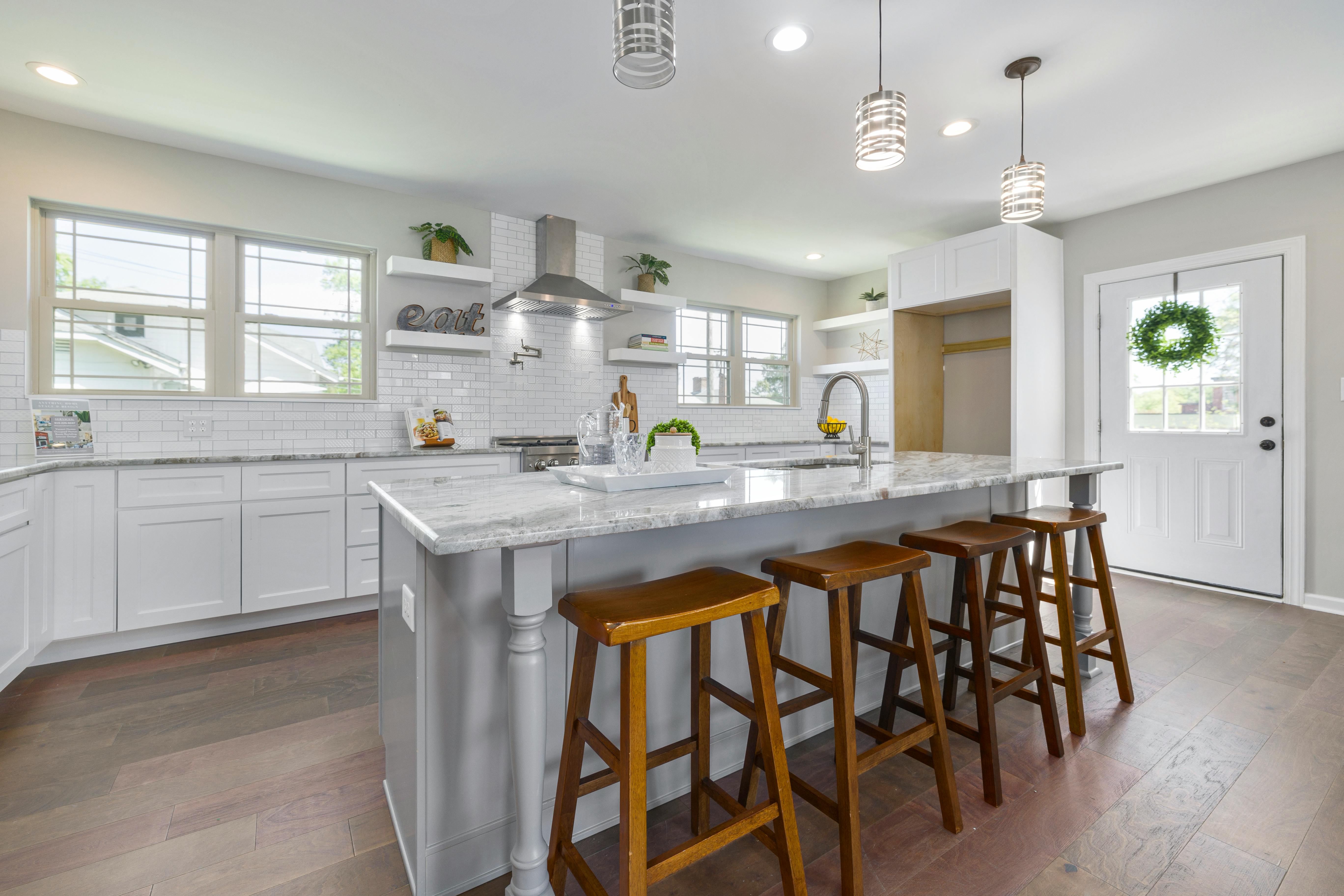 Modern white kitchen with a large marble island, four wooden bar stools, stainless steel range hood, and a door with a green wreath.