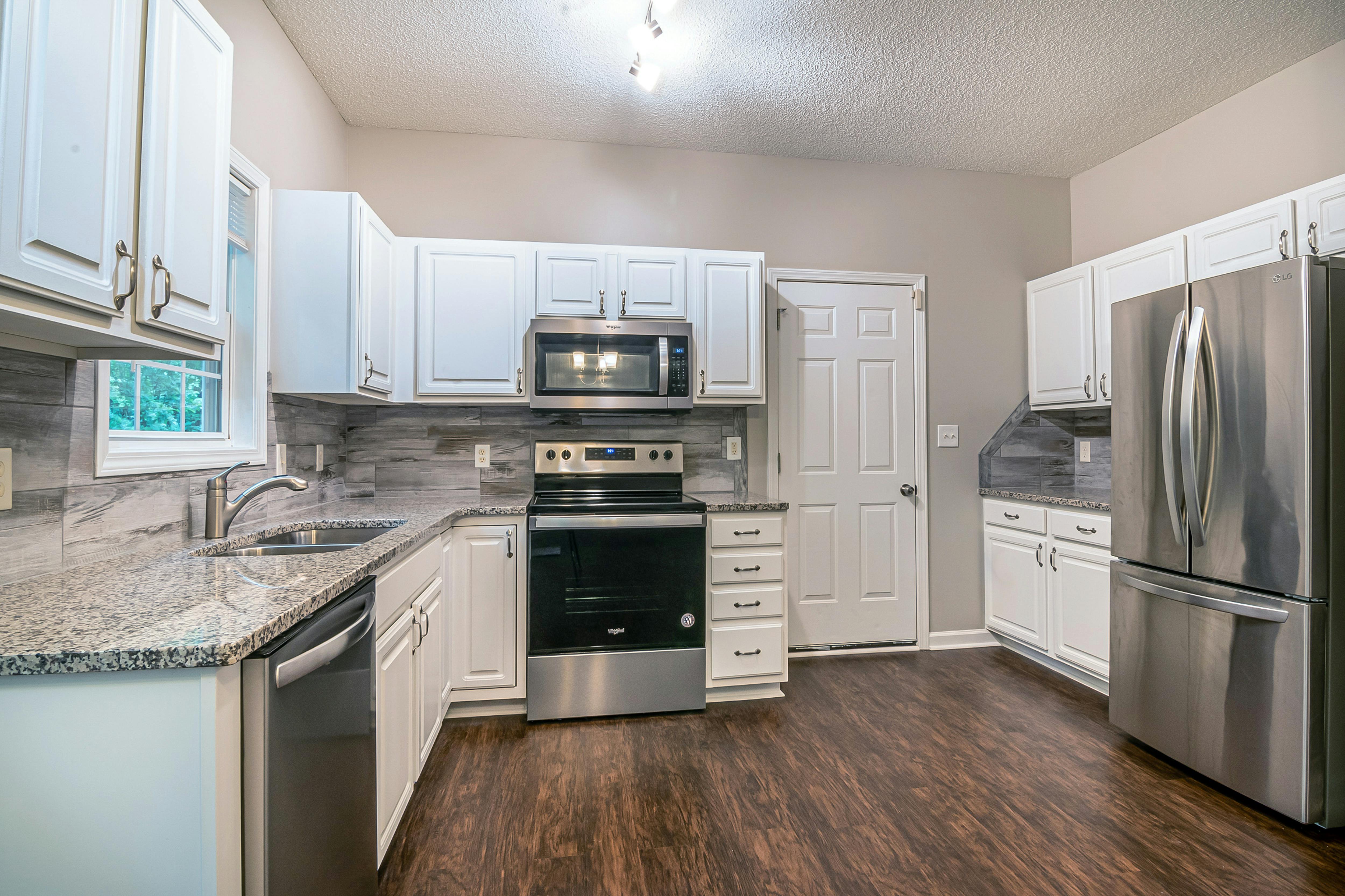 Modern kitchen with white cabinets, stainless steel appliances including refrigerator, oven, microwave, and dishwasher, granite countertops, and wooden flooring.