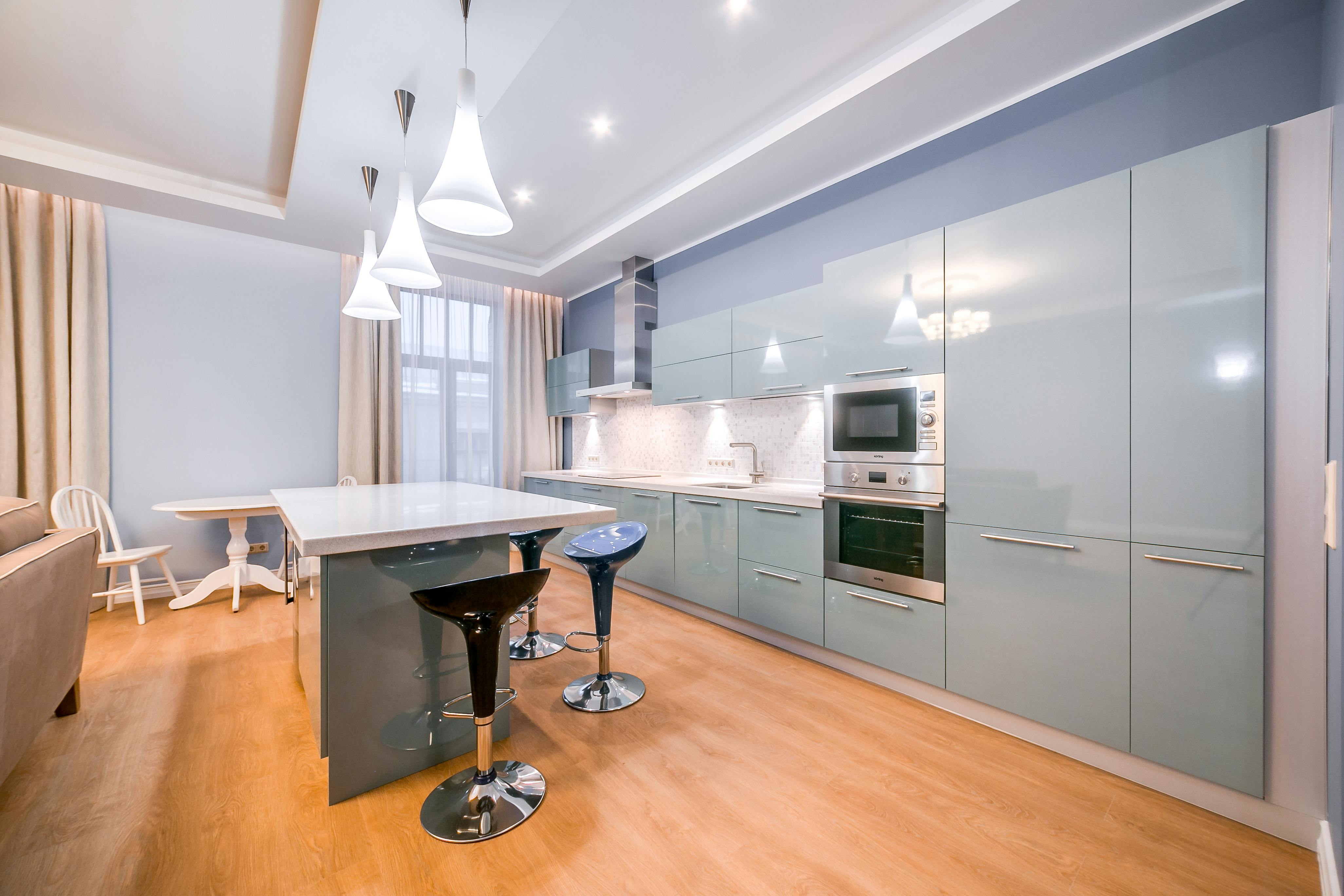Modern kitchen with glossy light blue cabinets, built-in oven and microwave, island with three bar stools, and pendant lights.