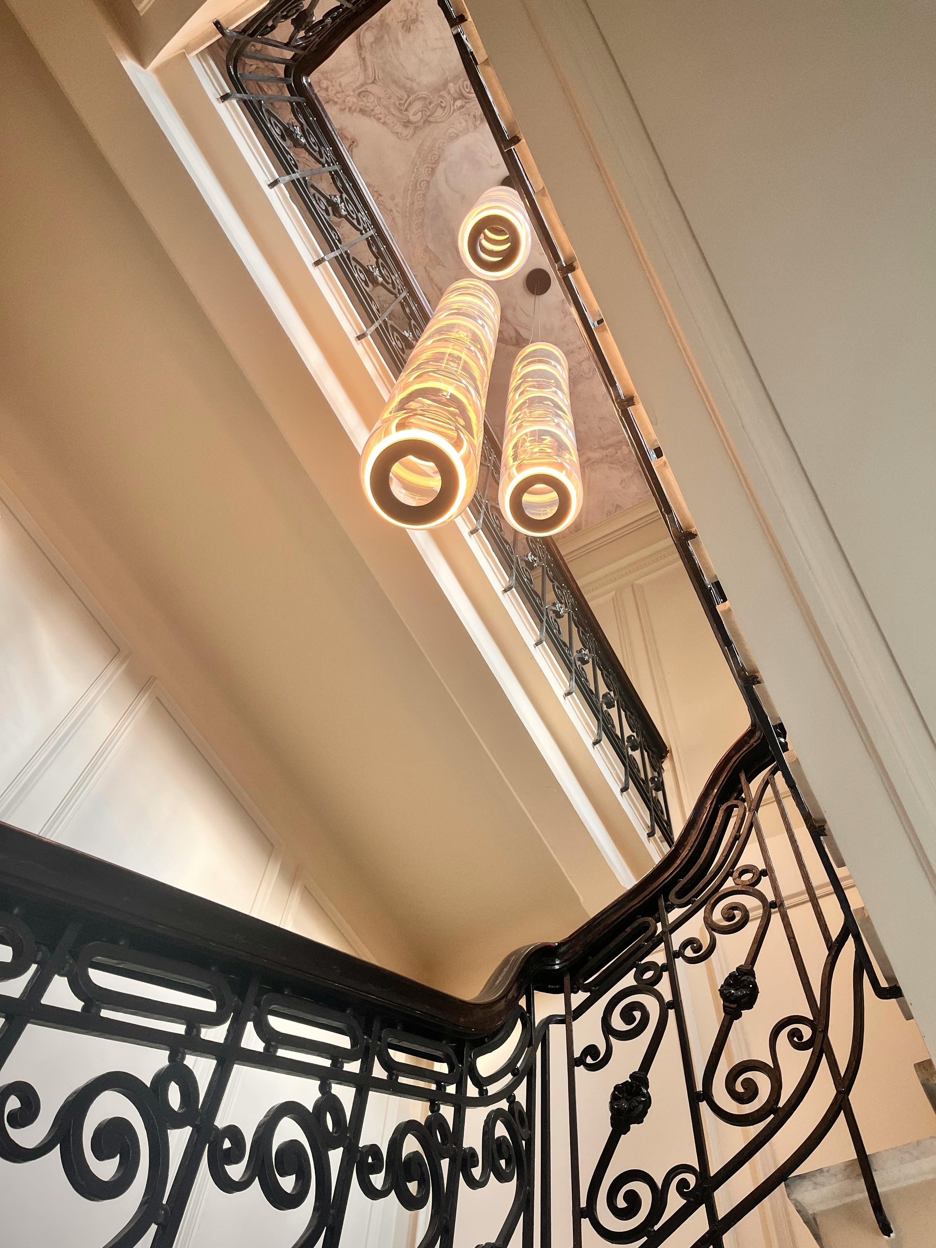 Modern cylindrical pendant lights hanging in a stairwell with ornate black wrought-iron railings and a decorative ceiling.