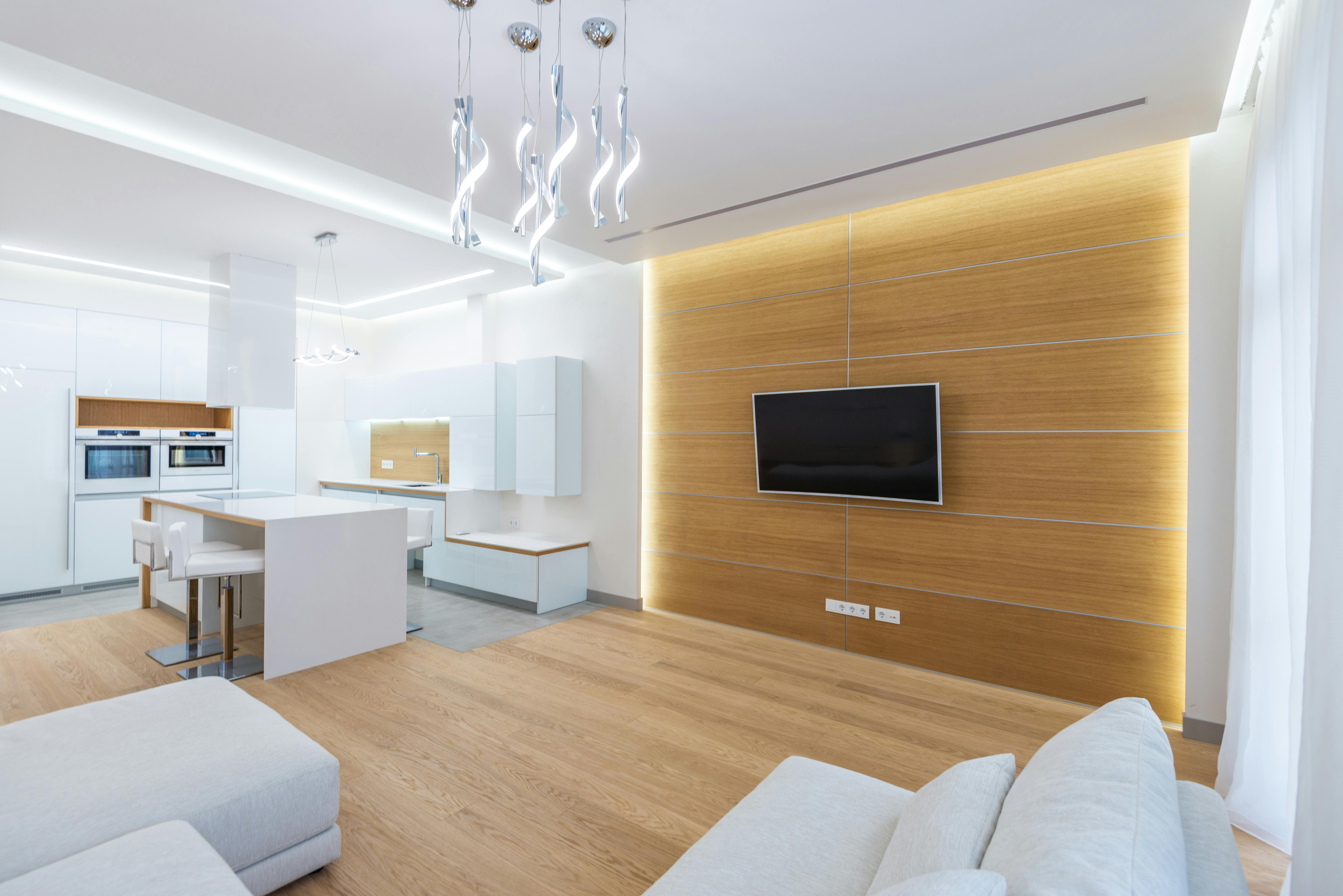 Modern open-plan living room and kitchen with white furniture, wooden flooring, and a wall-mounted flat-screen TV.