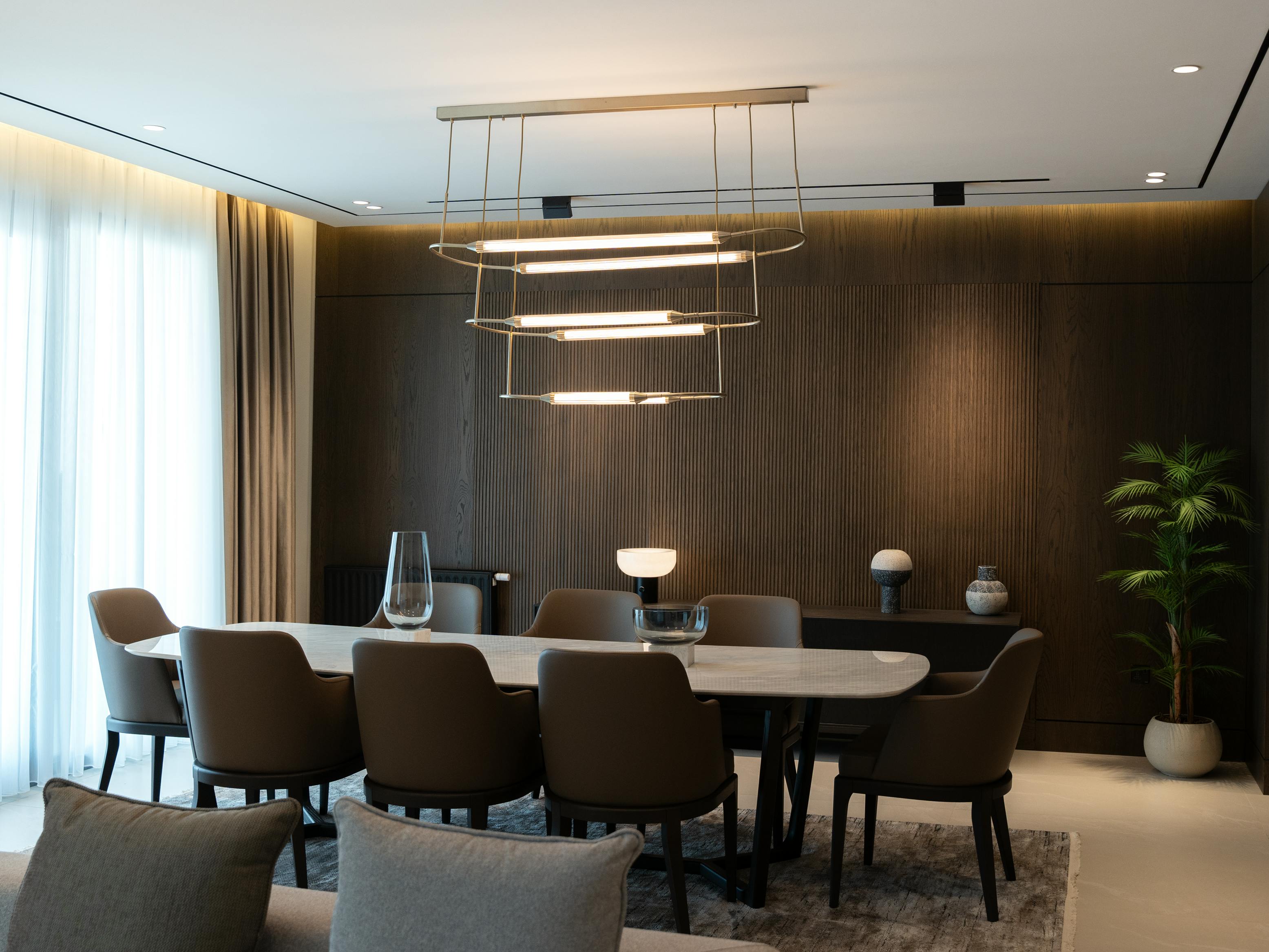 Modern dining room with a white oval table, eight brown chairs, ring-shaped pendant lights, and a wooden accent wall with decorative vases and a potted plant.