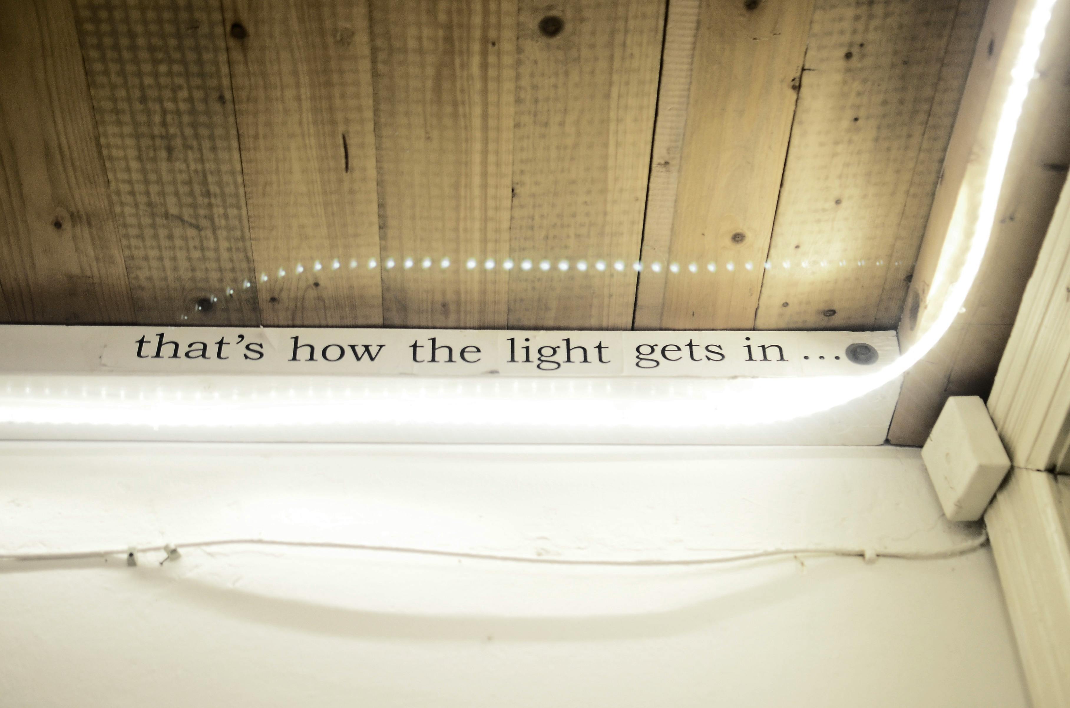 Wood ceiling panel with the phrase 'that's how the light gets in...' and white LED strip lighting along the edge.