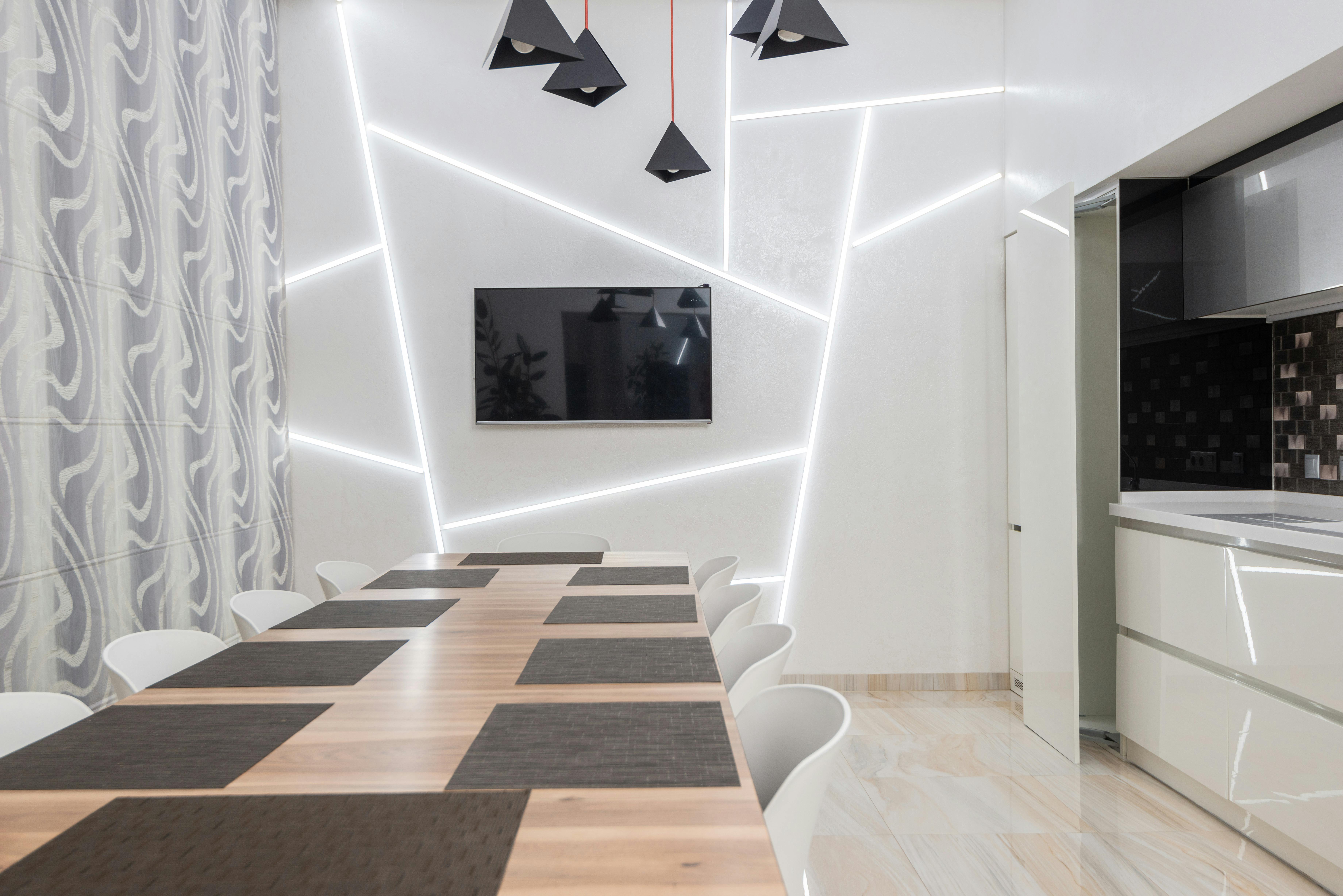 Modern dining room with a long wooden table set with dark placemats, white chairs, illuminated geometric wall designs, a mounted TV, and a partial view of a sleek kitchen.