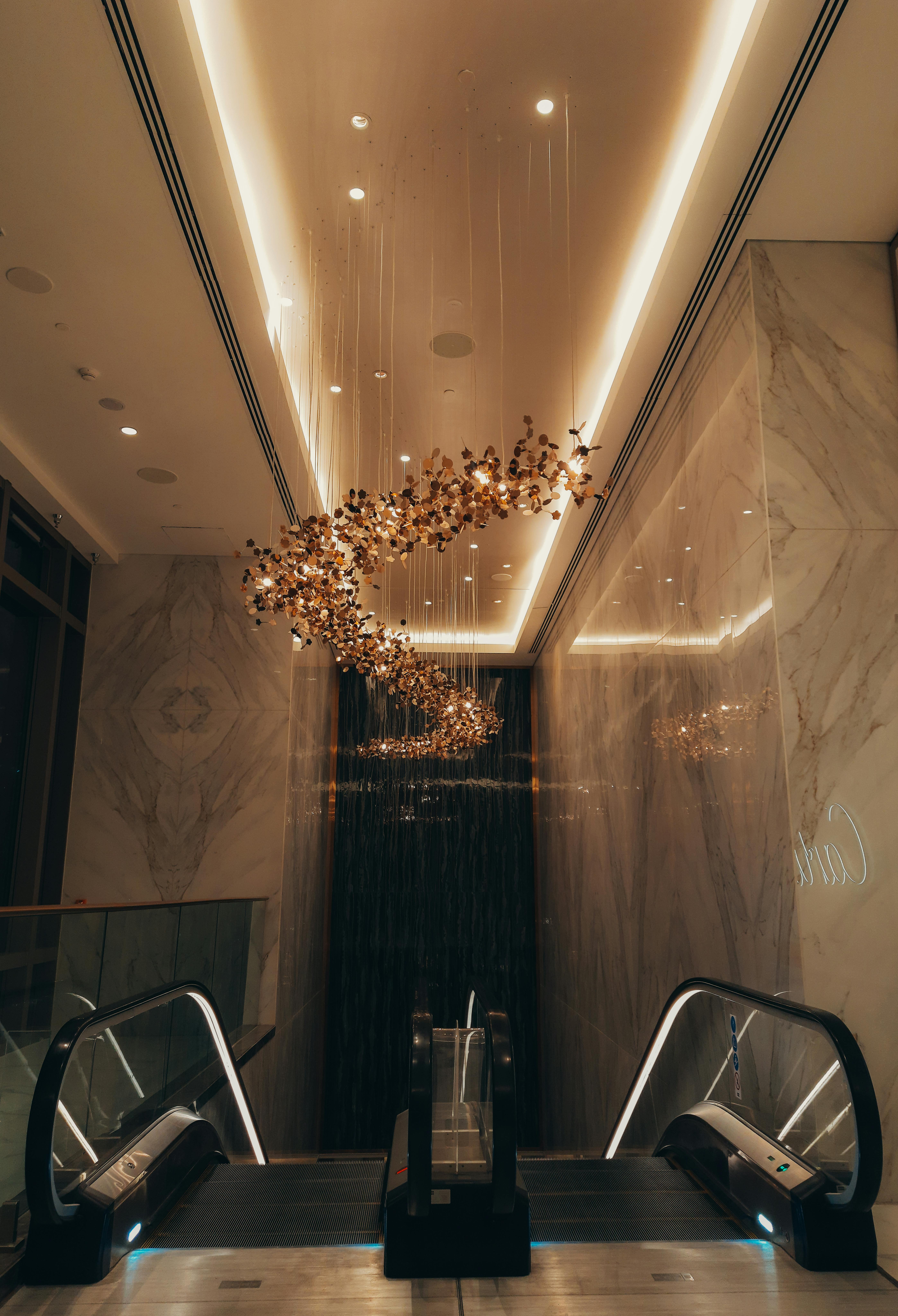 Upscale indoor escalators flanked by marble walls and a modern, abstract chandelier hanging from the ceiling.
