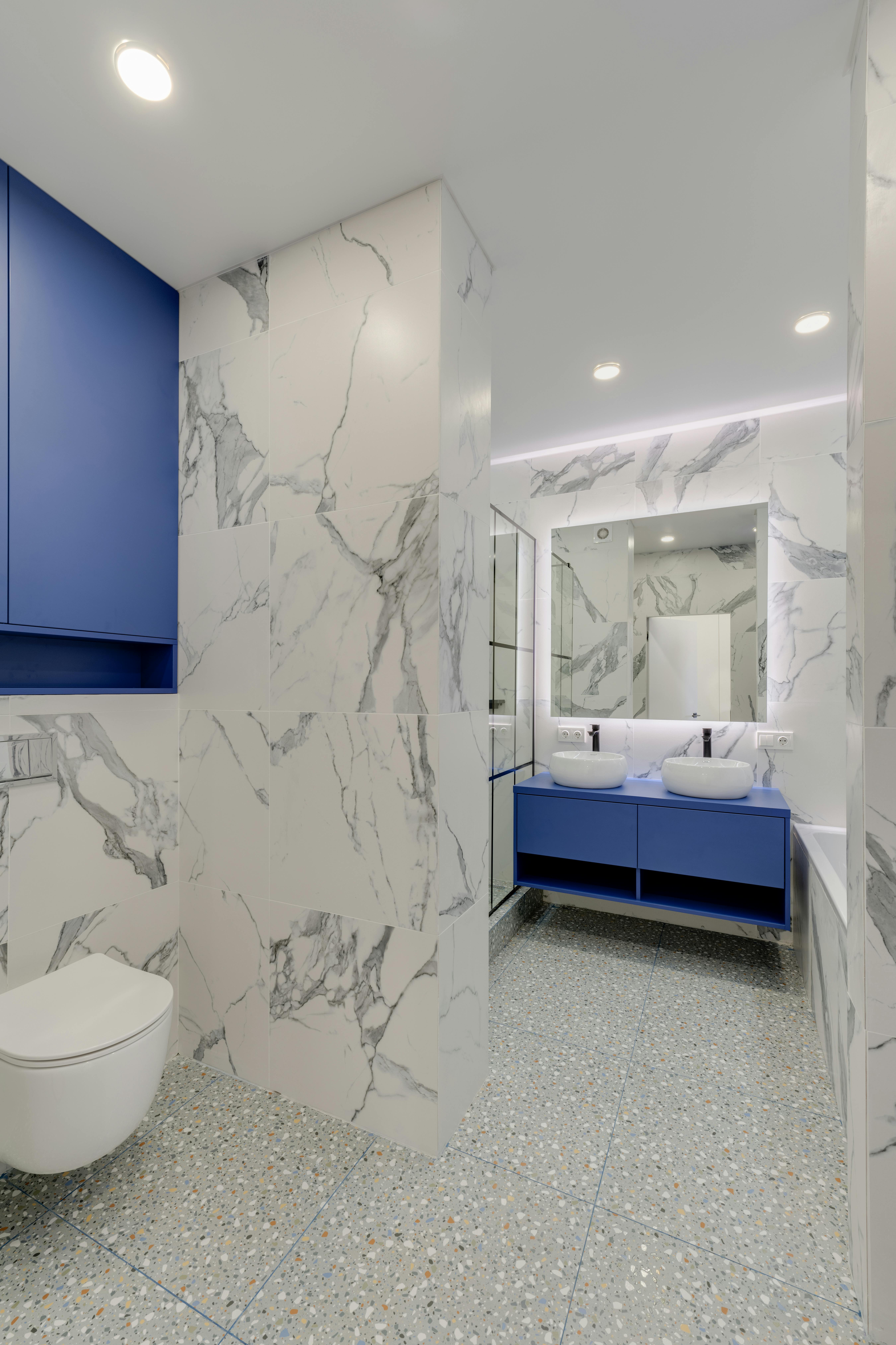 Modern bathroom with white marble-patterned walls, terrazzo floor, blue cabinets, double vessel sinks, and a wall-mounted toilet.