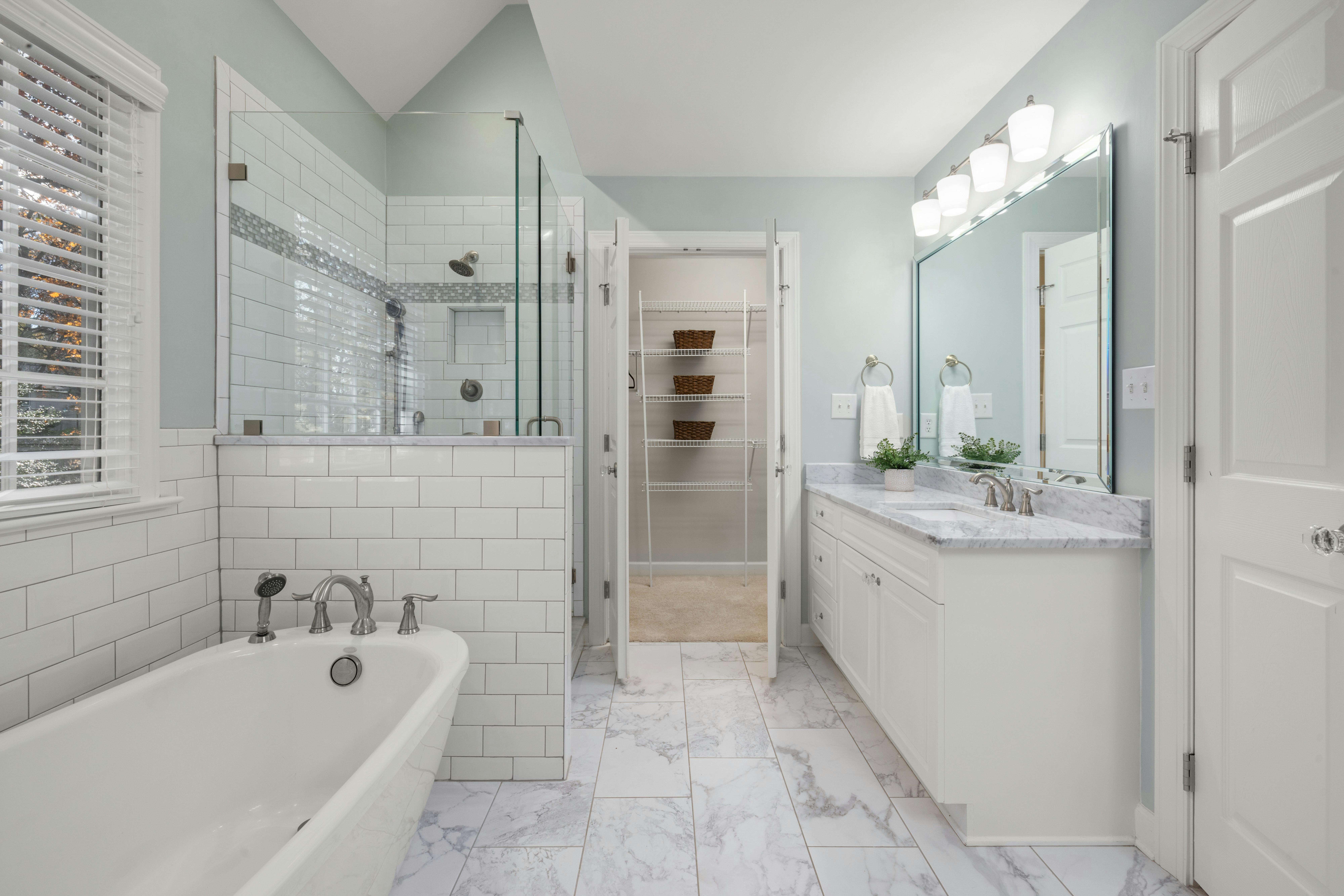Modern bathroom with a white bathtub, glass-enclosed shower, marble floor tiles, double vanity with marble countertop, and a doorway leading to a closet.