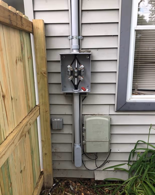 Outdoor electrical meter box with an open junction box above it, mounted on a house wall next to a wooden fence and a window.