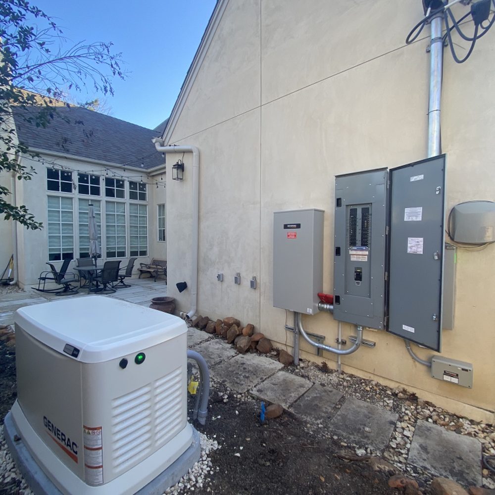 Outdoor Generac standby generator and electrical panel mounted on the exterior wall of a beige house with a patio and chairs in the background.