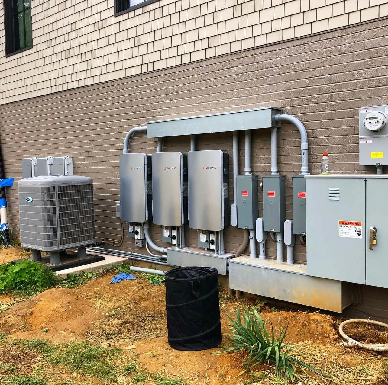 Outdoor electrical setup mounted on a brick wall including three Enphase inverters, electrical panels, a large air conditioning unit, and a Gatorade bottle on top of one panel.