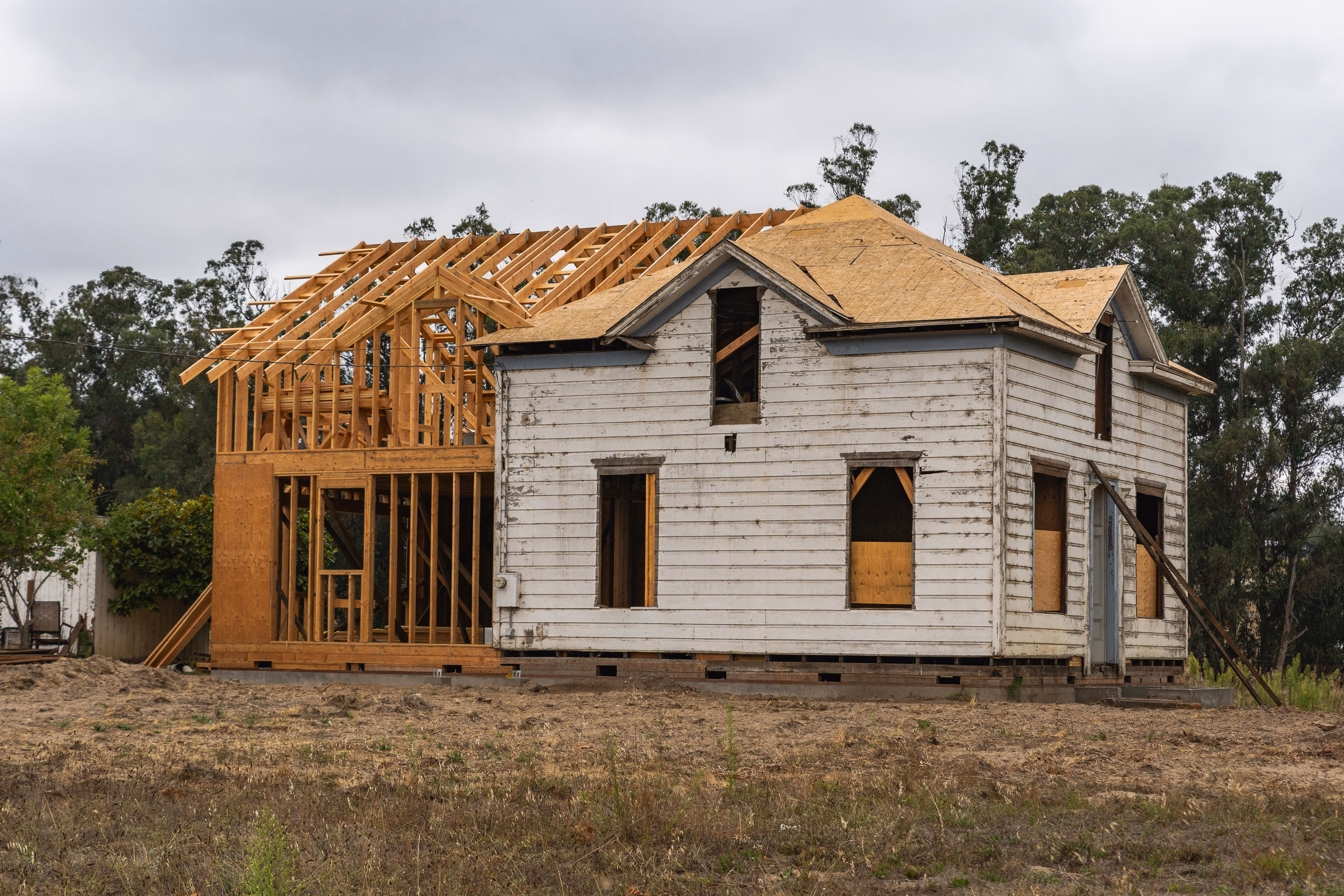 Old white wooden house undergoing renovation with new wooden framework and roof structure being added.