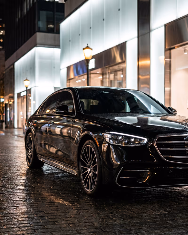 Black luxury sedan parked on a wet cobblestone street at night with illuminated modern buildings.