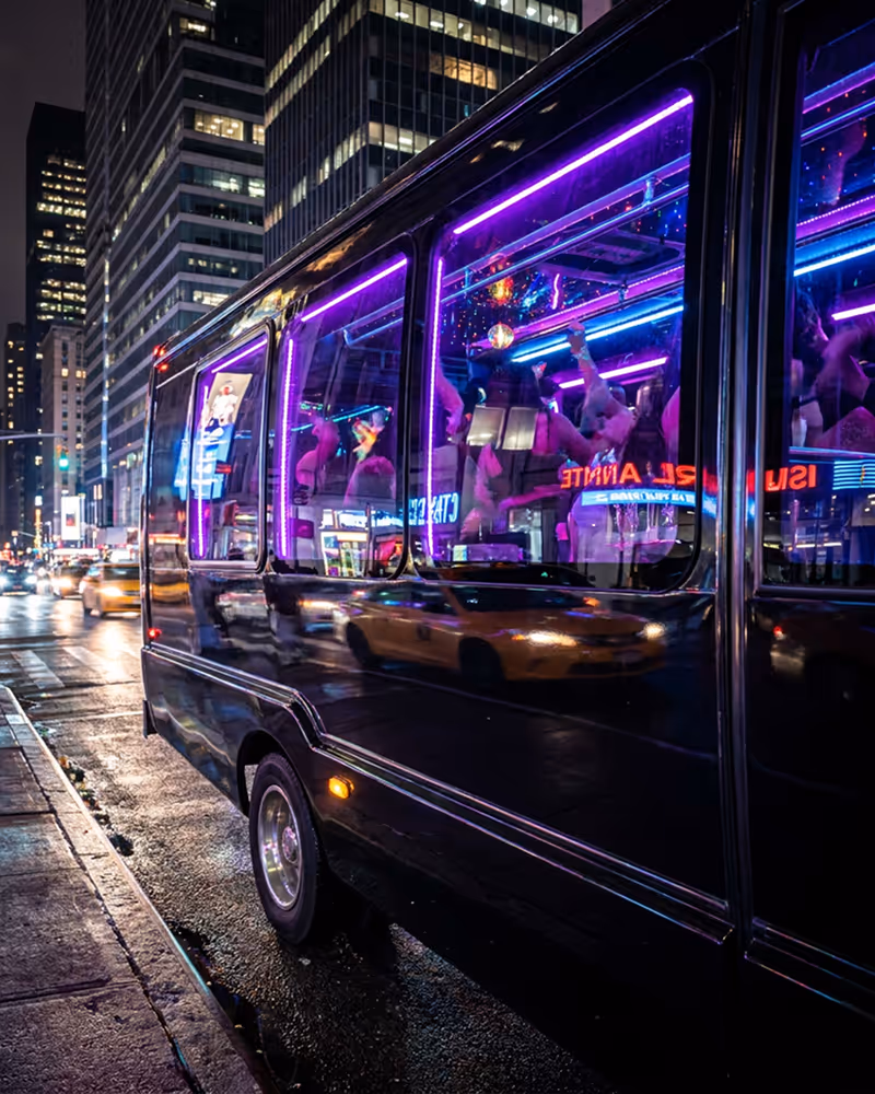 Black party bus with purple neon lights parked on a city street at night reflecting taxis and buildings.
