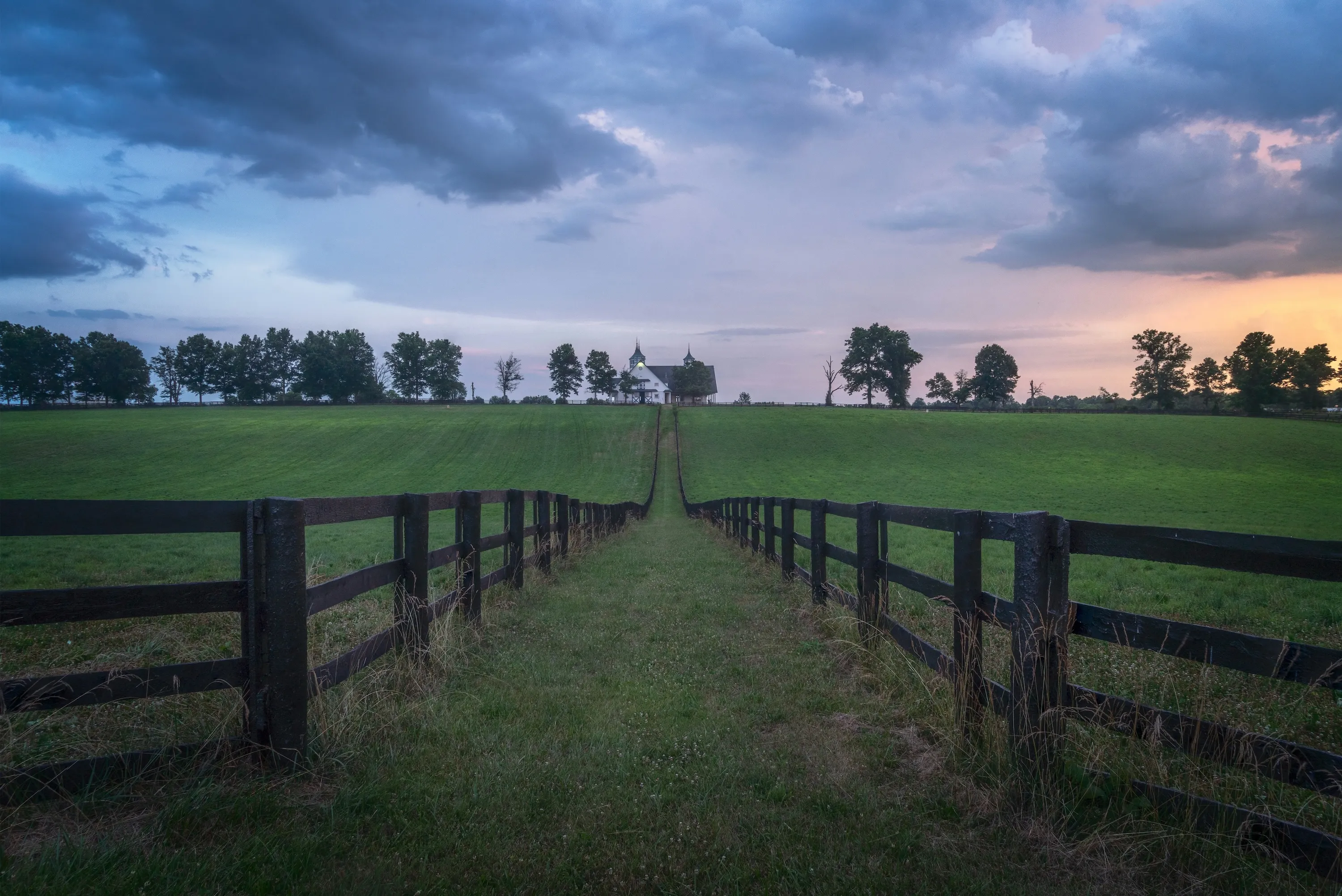 Long wooden fence lines a grassy path leading to a white farmhouse on a hill under a cloudy sky at dusk.