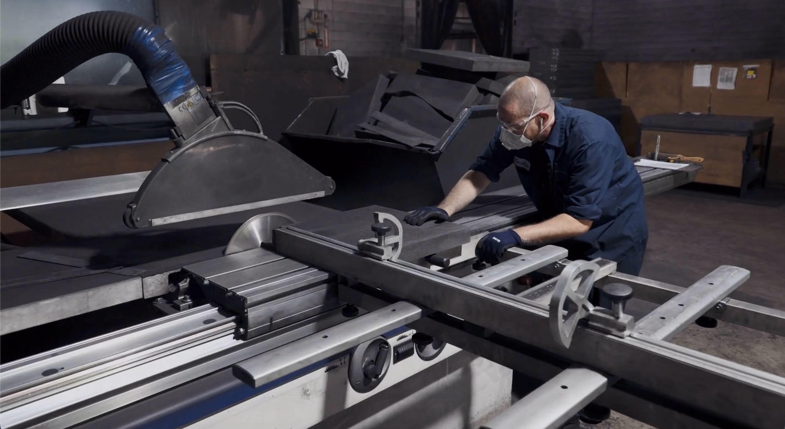 Worker in safety gloves and mask operating a large industrial cutting saw on a foam sheet.