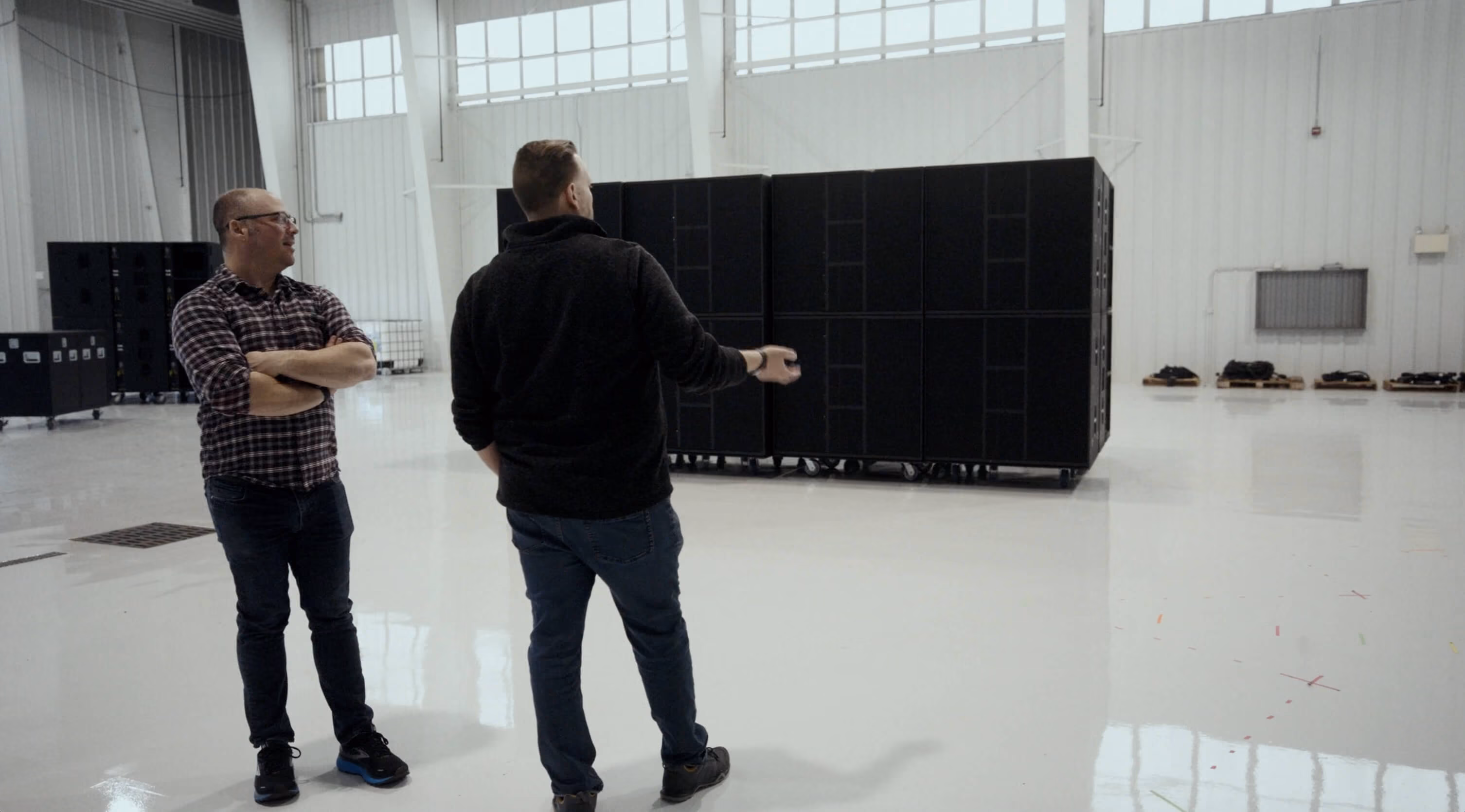 Two men in a large bright room, one gesturing toward a stack of black server racks on wheels.