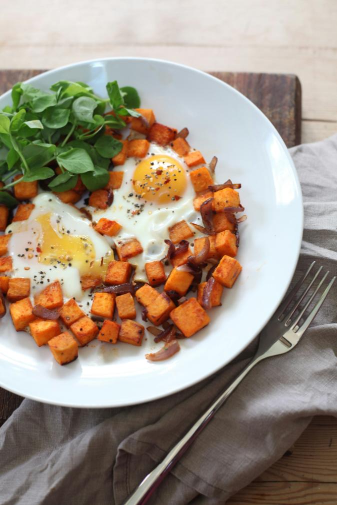 Sweet potato hash in a cast iron skillet with crispy sweet potato cubes, red pepper, kale and four fried eggs with runny yolks