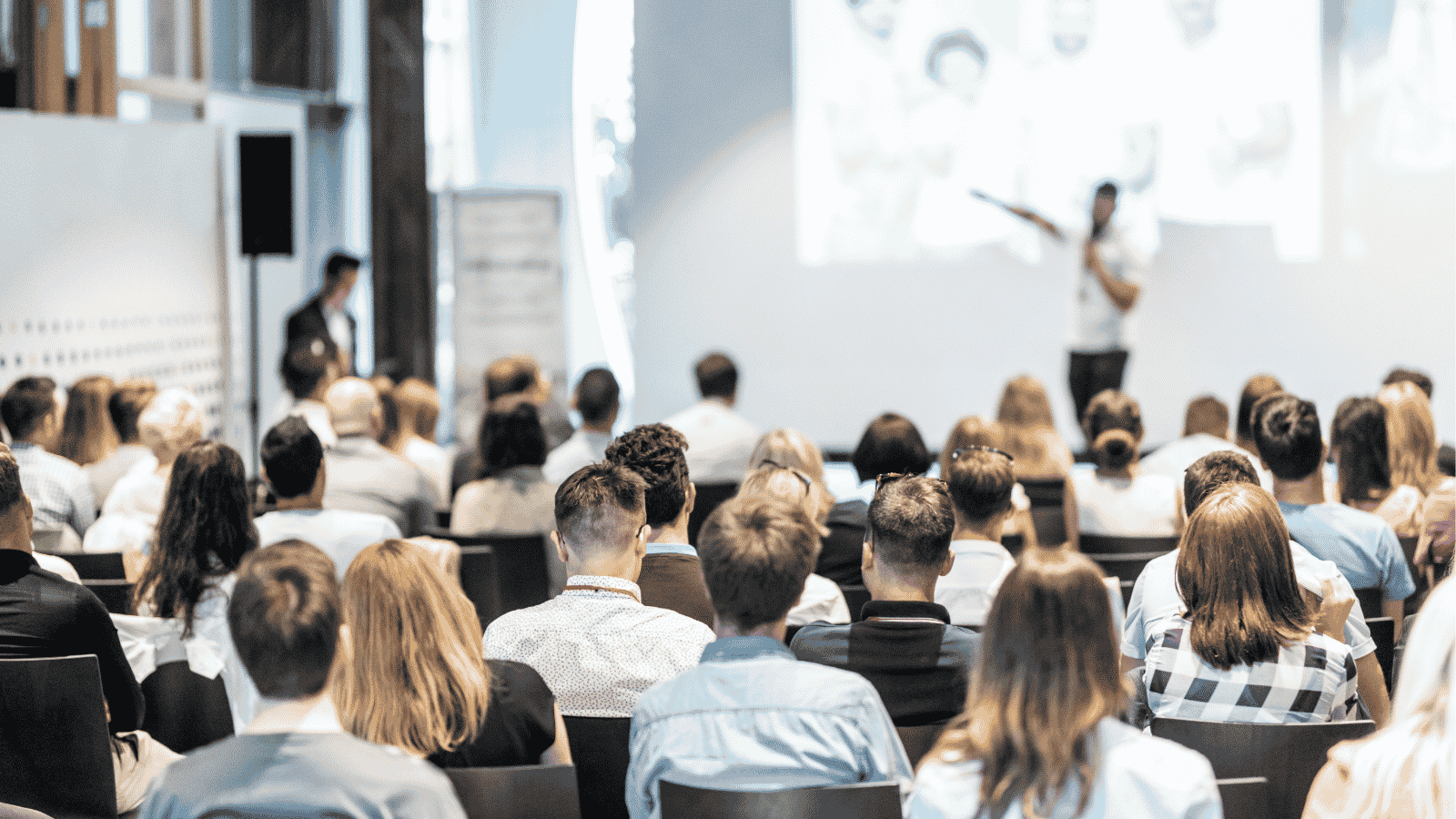 Audience sitting in rows watching a speaker presenting in front of a screen in a bright room.