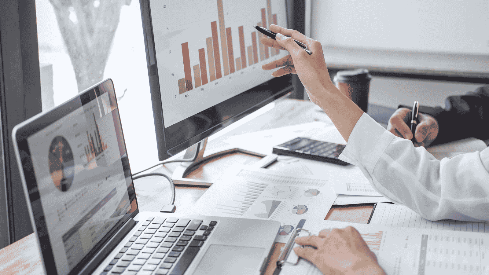 Two people analyzing bar and pie charts on laptop and desktop screens at a desk with documents.