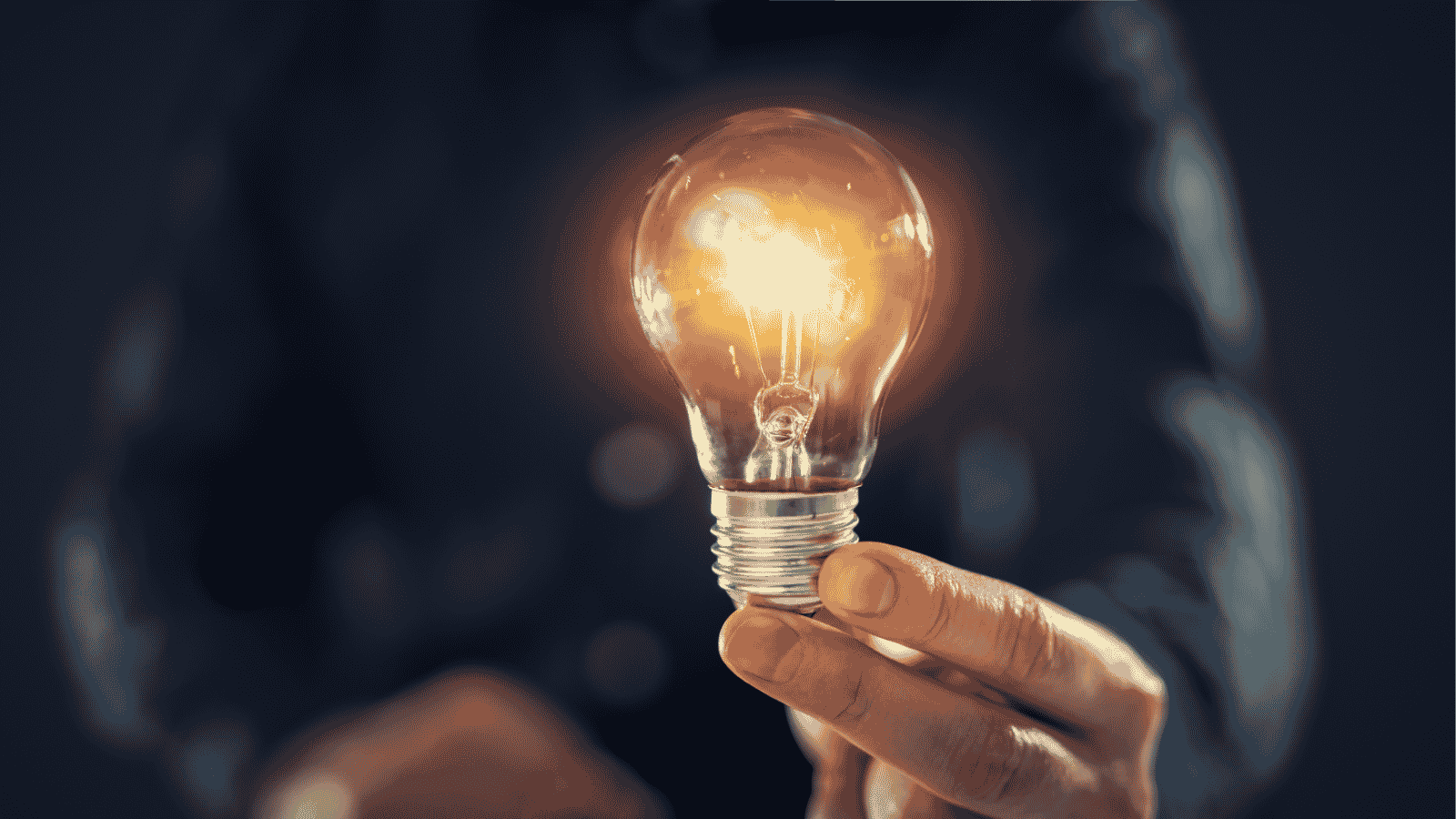 Hand holding a glowing incandescent light bulb with a dark, blurred background.