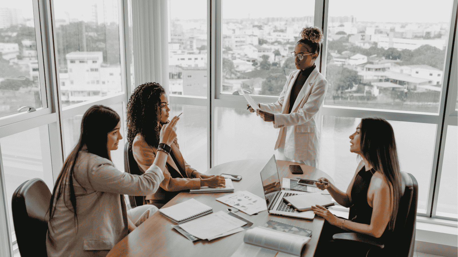 Four professional women in a meeting room with city views, discussing and reviewing documents.