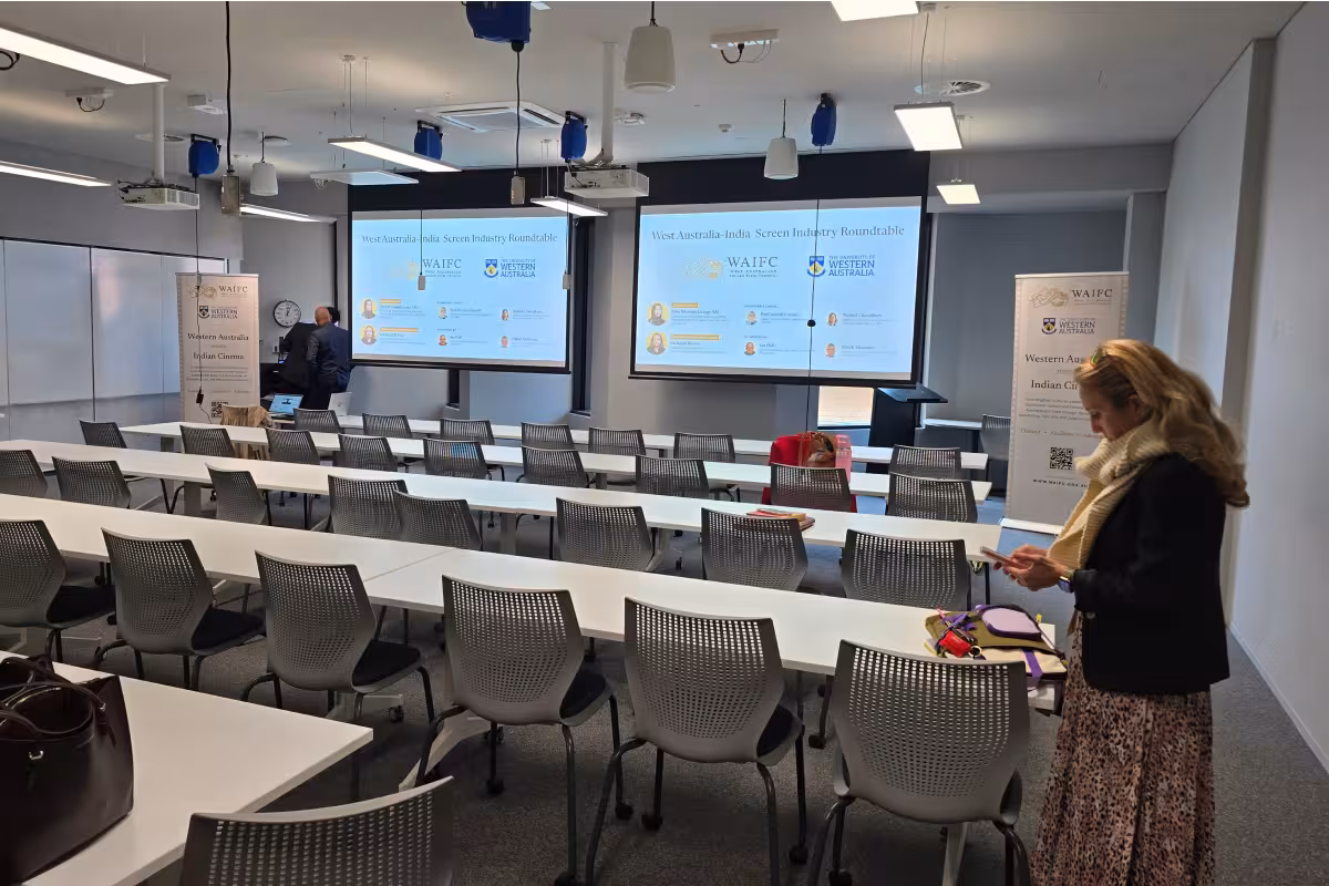 Conference room with rows of chairs and two screens showing West Australia-India Screen Industry Roundtable.