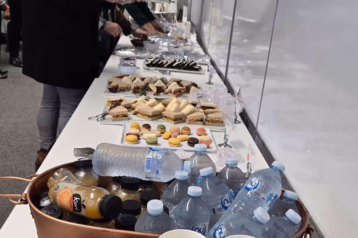 A buffet table with water bottles, orange juice bottles, macarons, sandwiches, and people serving themselves.