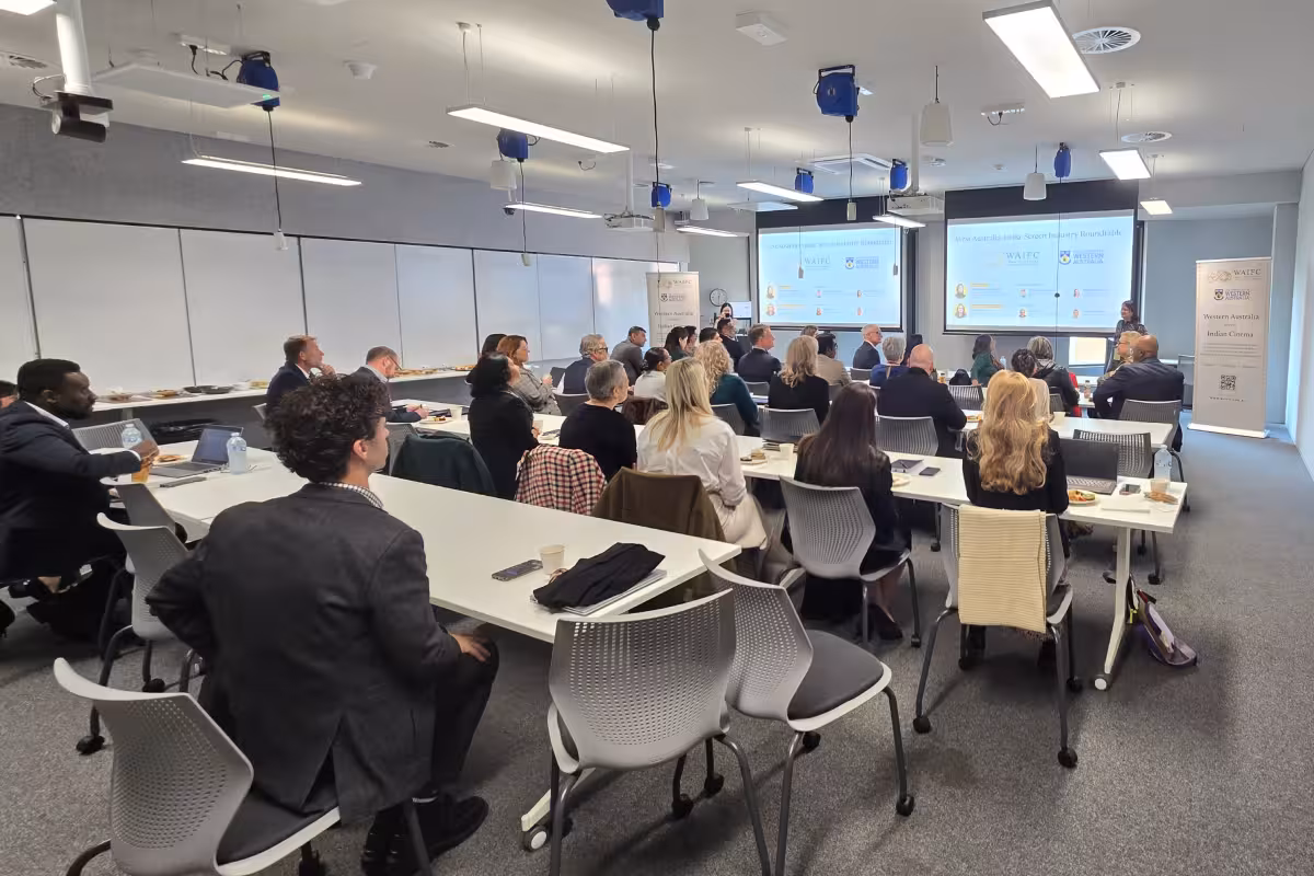 Group of people seated at tables in a conference room watching a presentation on two large screens.
