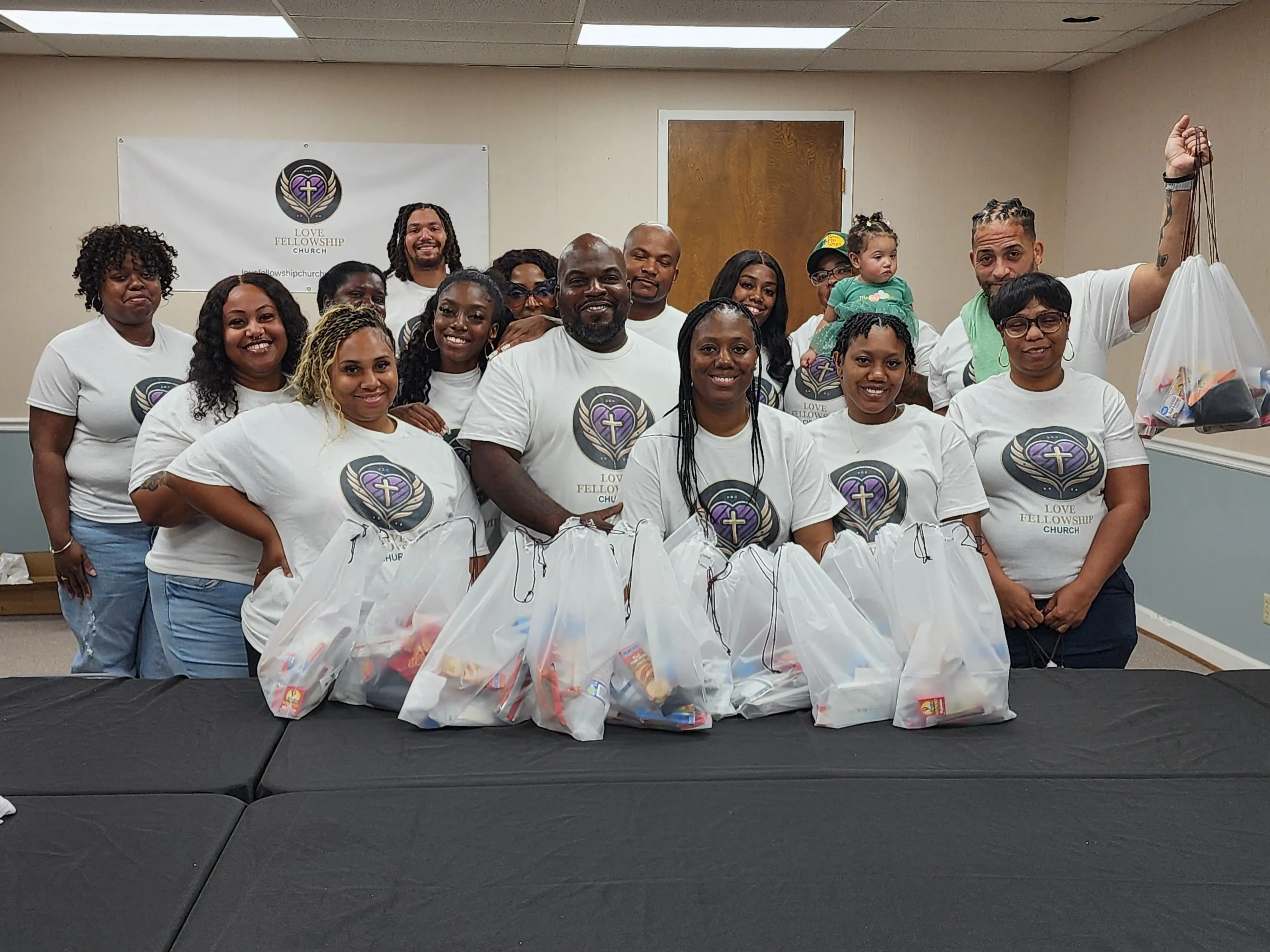 Group of people in matching shirts hold bags of food while posing in a room.