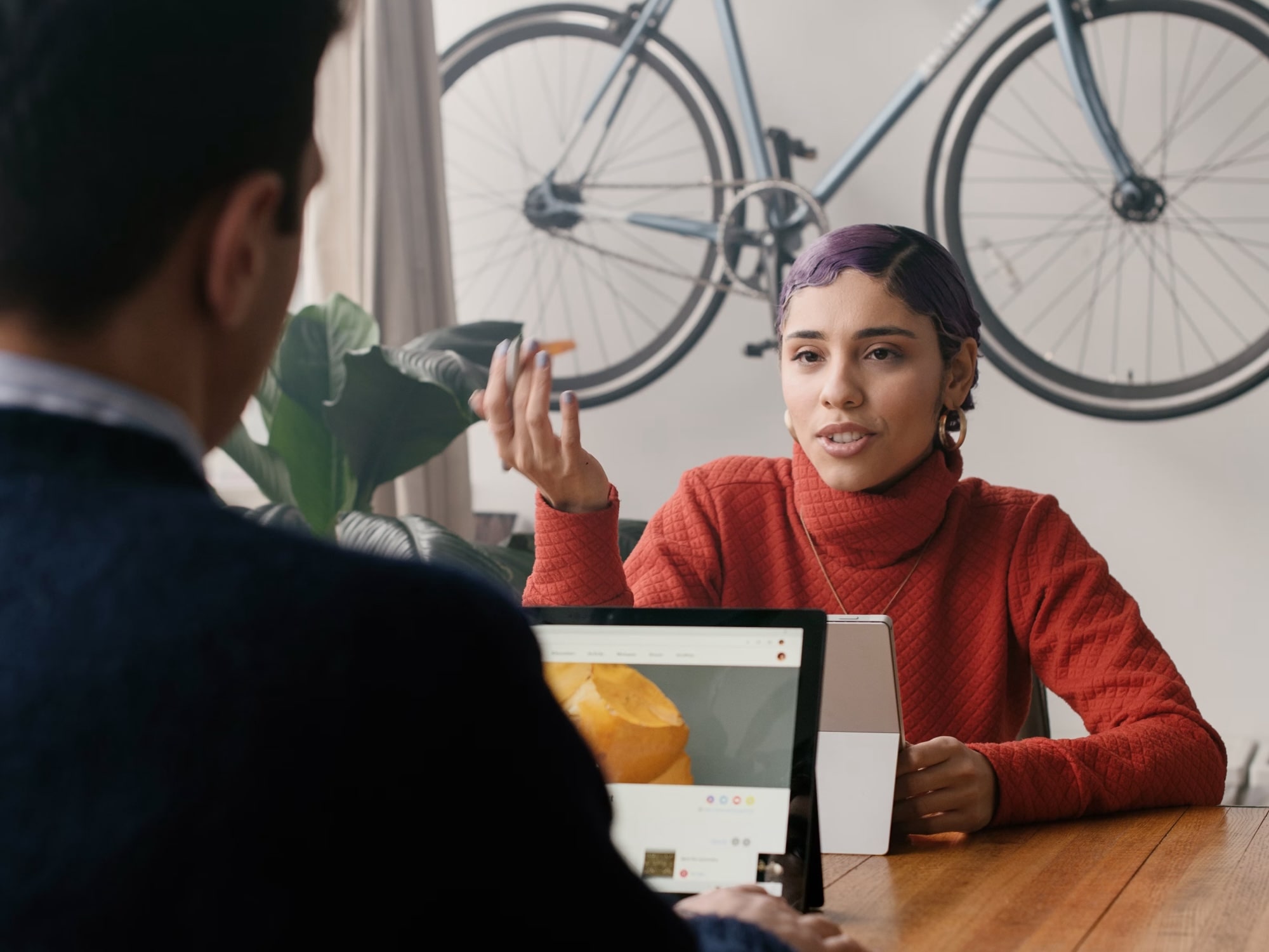 A woman in a red sweater talks to a man working on a tablet in a modern room with a bicycle mounted on the wall.
