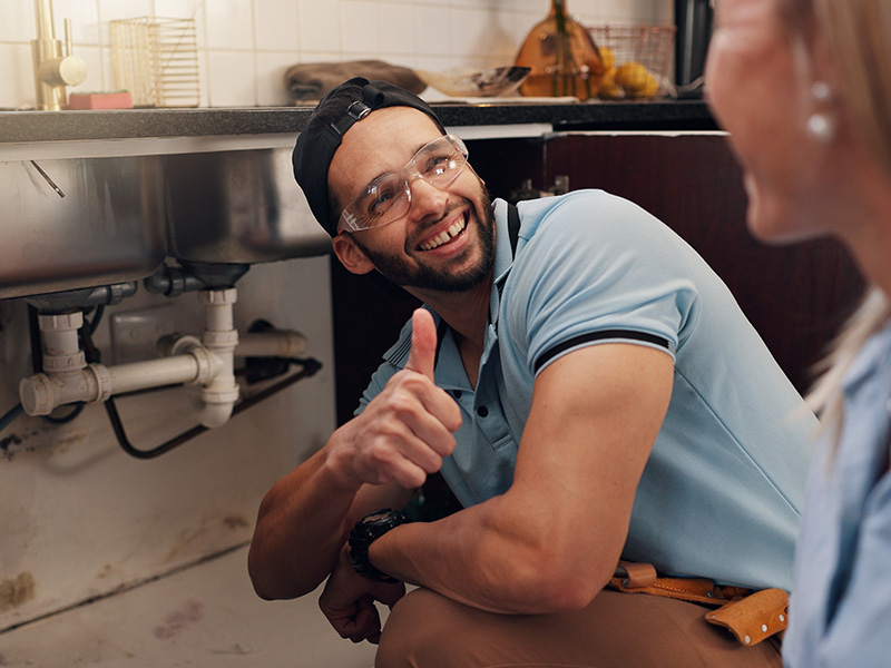 Smiling plumber wearing safety glasses giving a thumbs up while working under a kitchen sink.