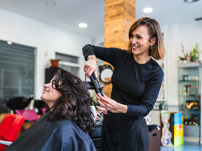 Hairdresser smiling while curling a woman's dark hair in a salon.