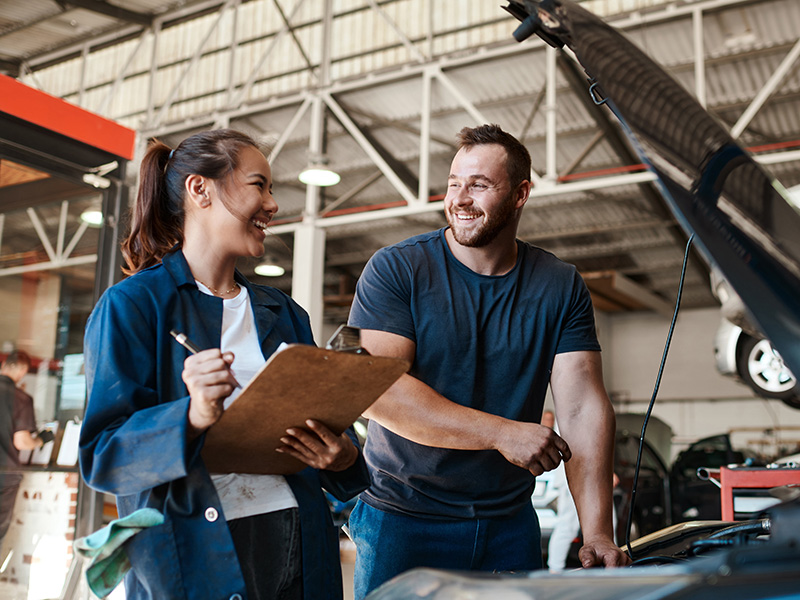 Two mechanics, a woman with a clipboard and a man leaning on an open car hood, smiling and talking in a garage.