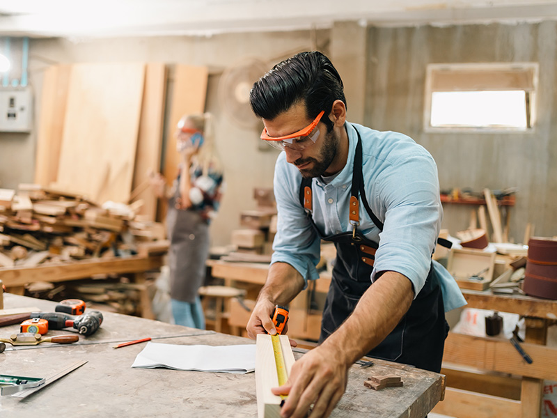 A male carpenter wearing orange safety glasses measures a wooden plank in a workshop with tools and wood in the background.