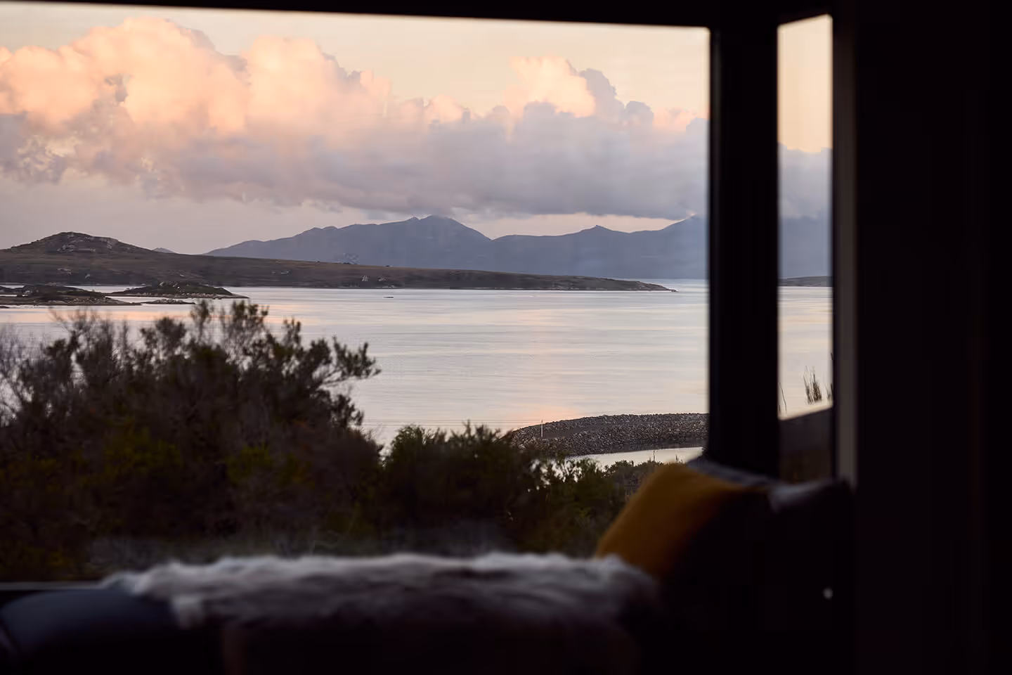 View of calm water, distant hills, and pink clouds seen through a window with blurred indoor seating.