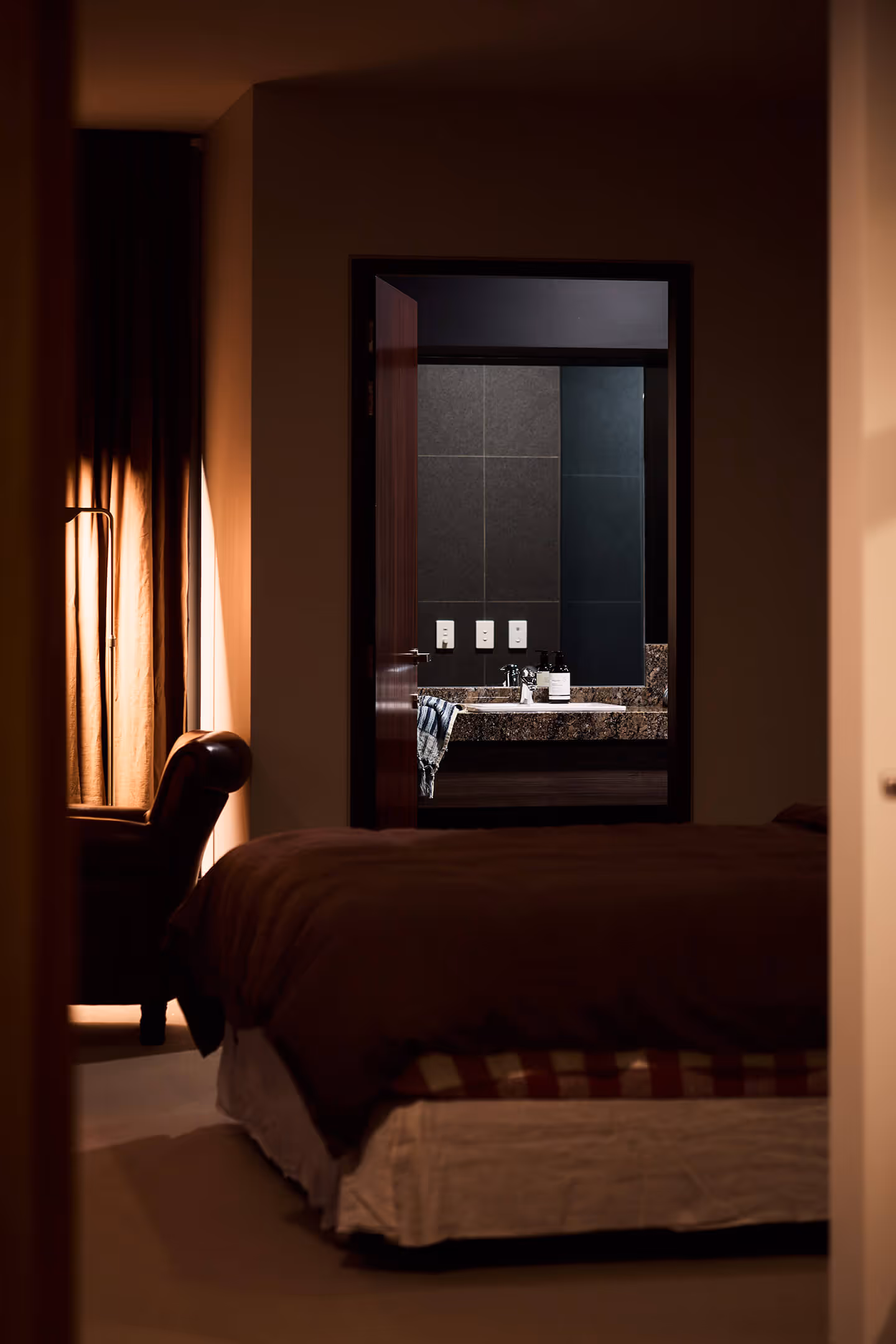 Dimly lit bedroom showing bed, armchair near curtained window, and open door to bathroom sink area.