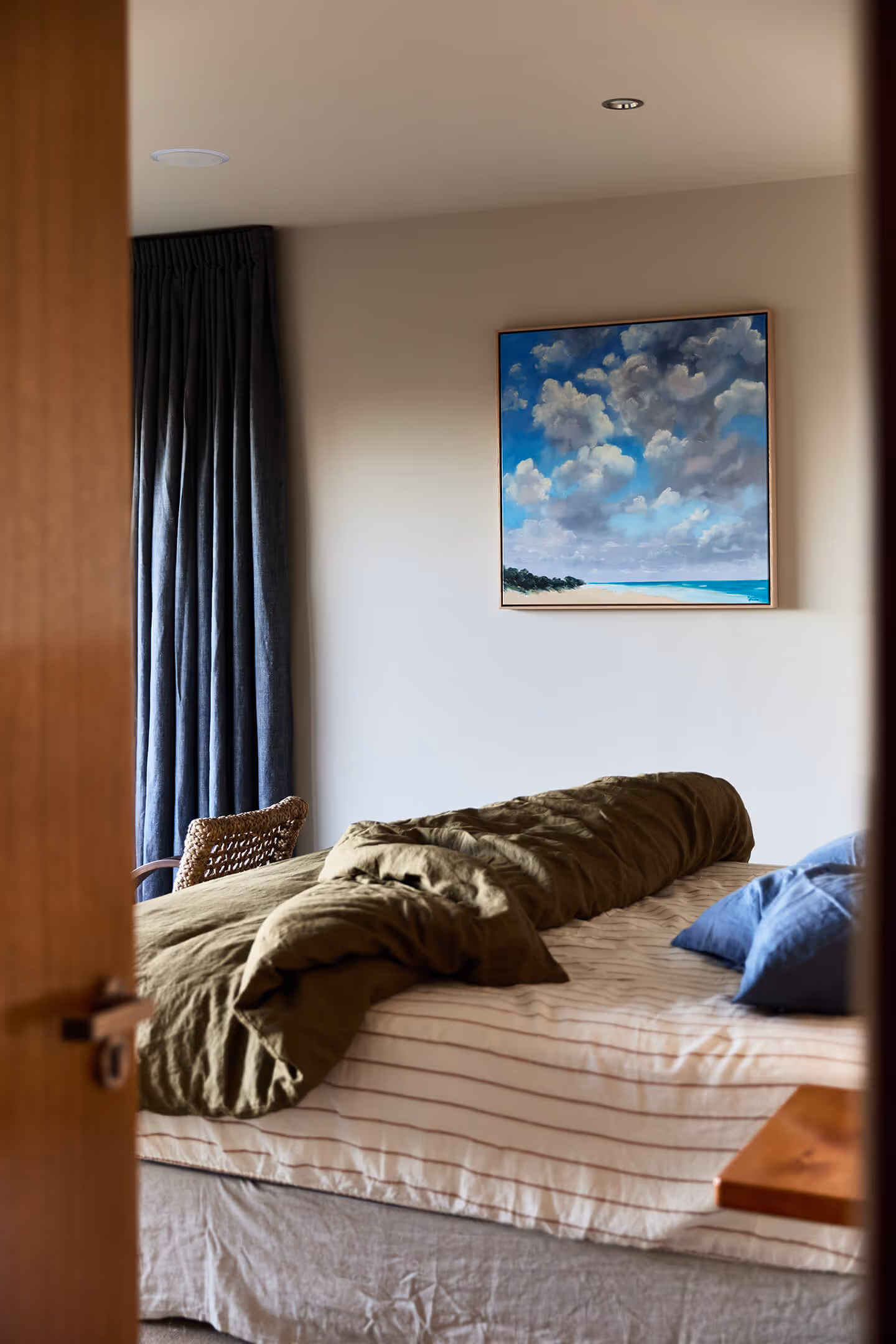 Bedroom with unmade bed, green blanket, blue pillows, woven chair, and ocean cloud painting on wall.
