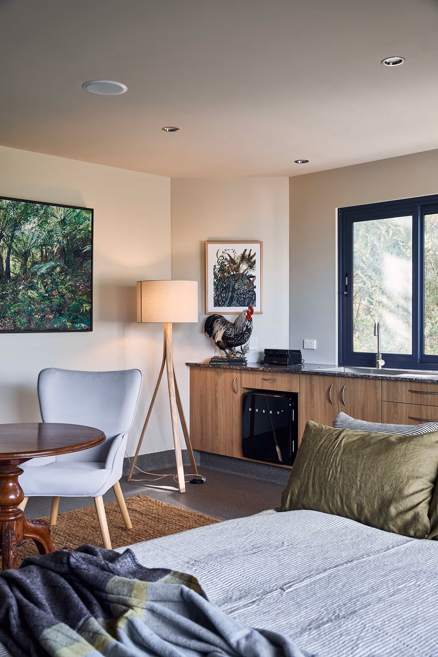 Cozy room with a bed, chair, round wooden table, floor lamp, kitchenette, and framed botanical art.