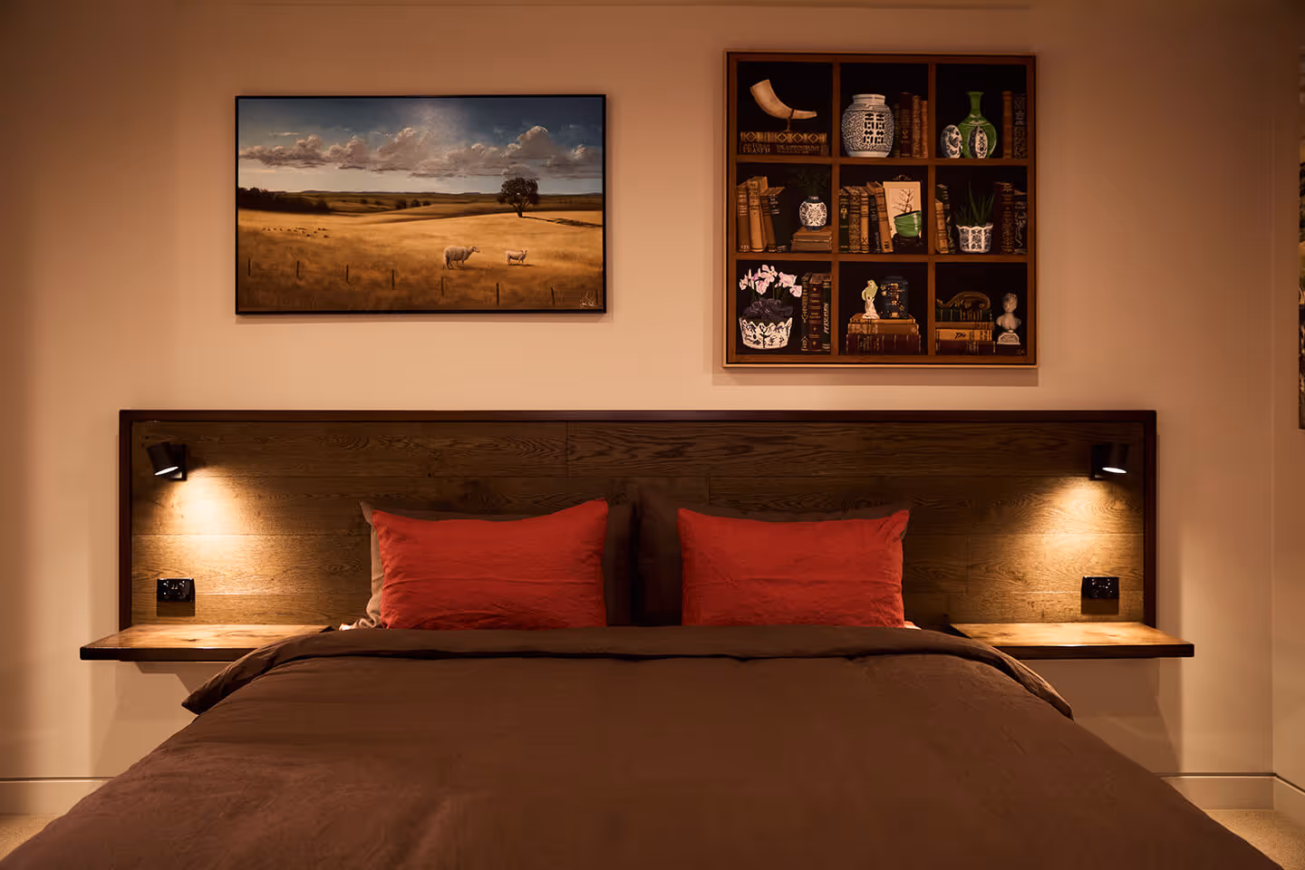 Bed with dark brown bedding, red pillows, wooden headboard, two wall lamps, and framed art above.