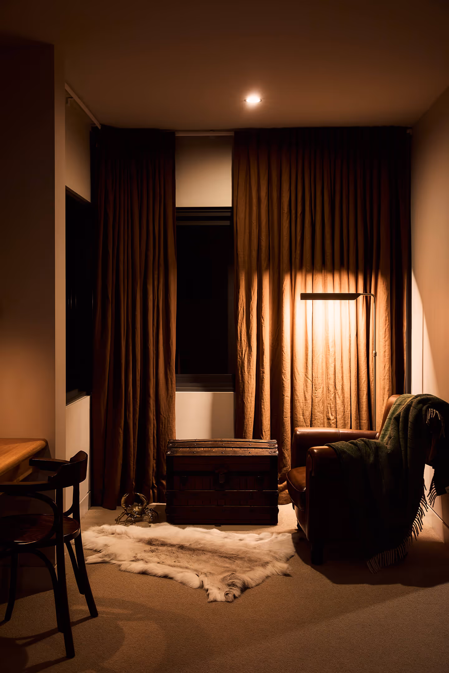 Cozy room with brown curtains, leather armchair with blanket, wooden chest, and soft rug under warm light.