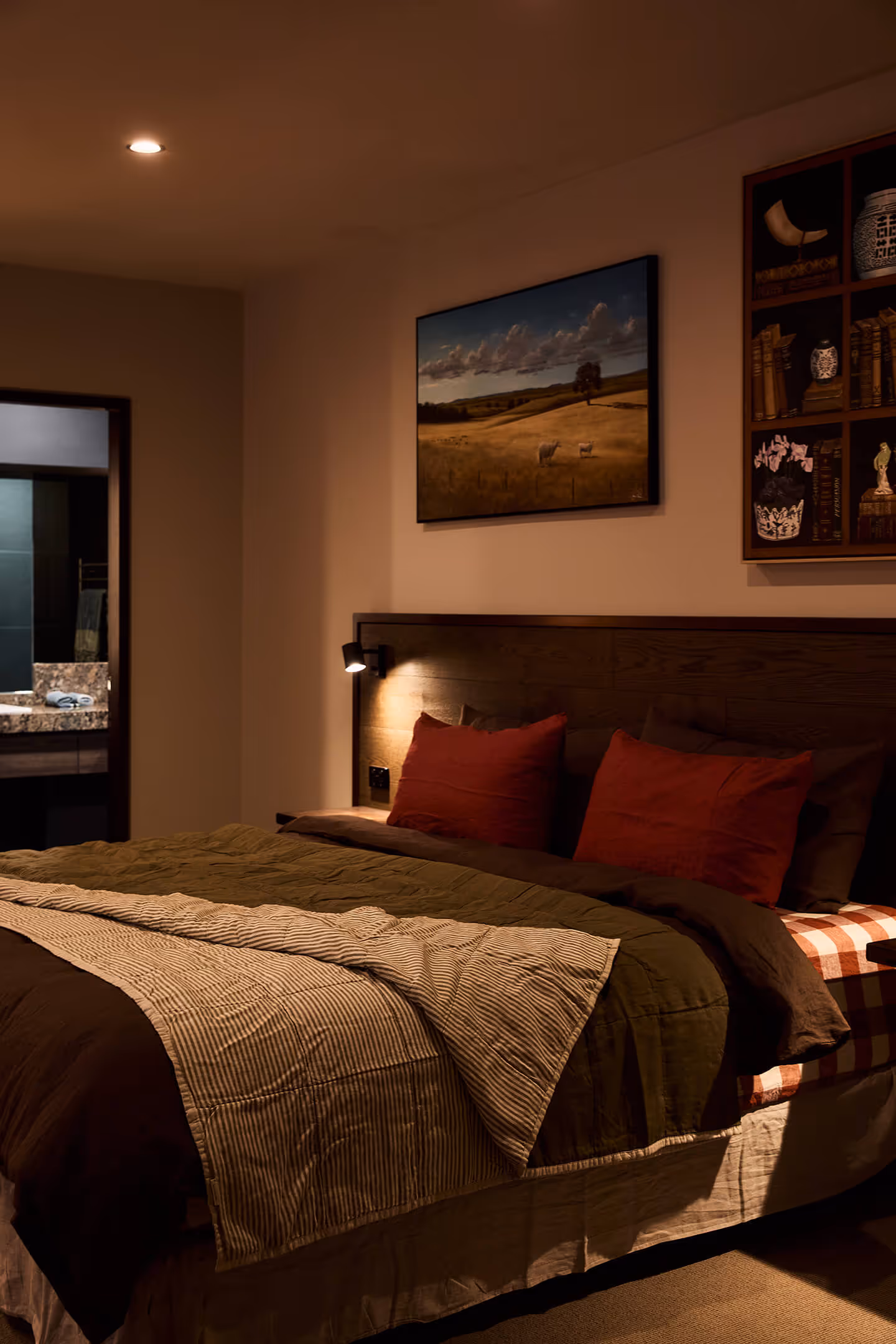 Cozy bedroom with layered bedding, red pillows, illuminated reading light, and landscape wall art.