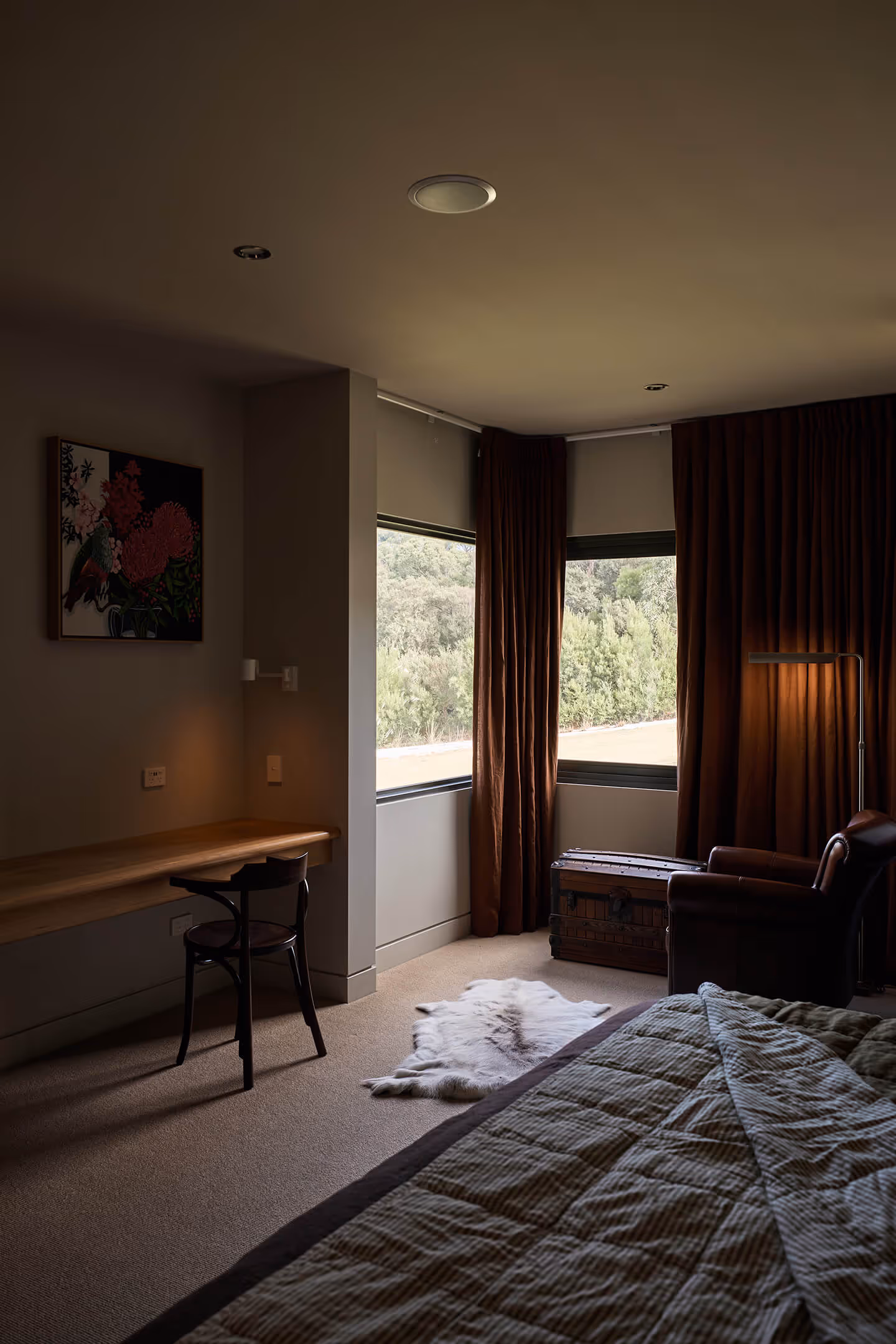 Cozy bedroom corner with a bed, leather armchair, wooden chest, desk with chair, and large windows with brown curtains.
