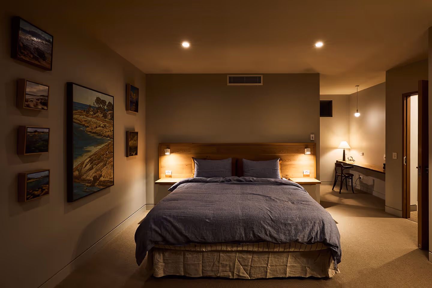 Cozy bedroom with a queen bed, blue bedding, wooden headboard, wall art, and a lit desk area.