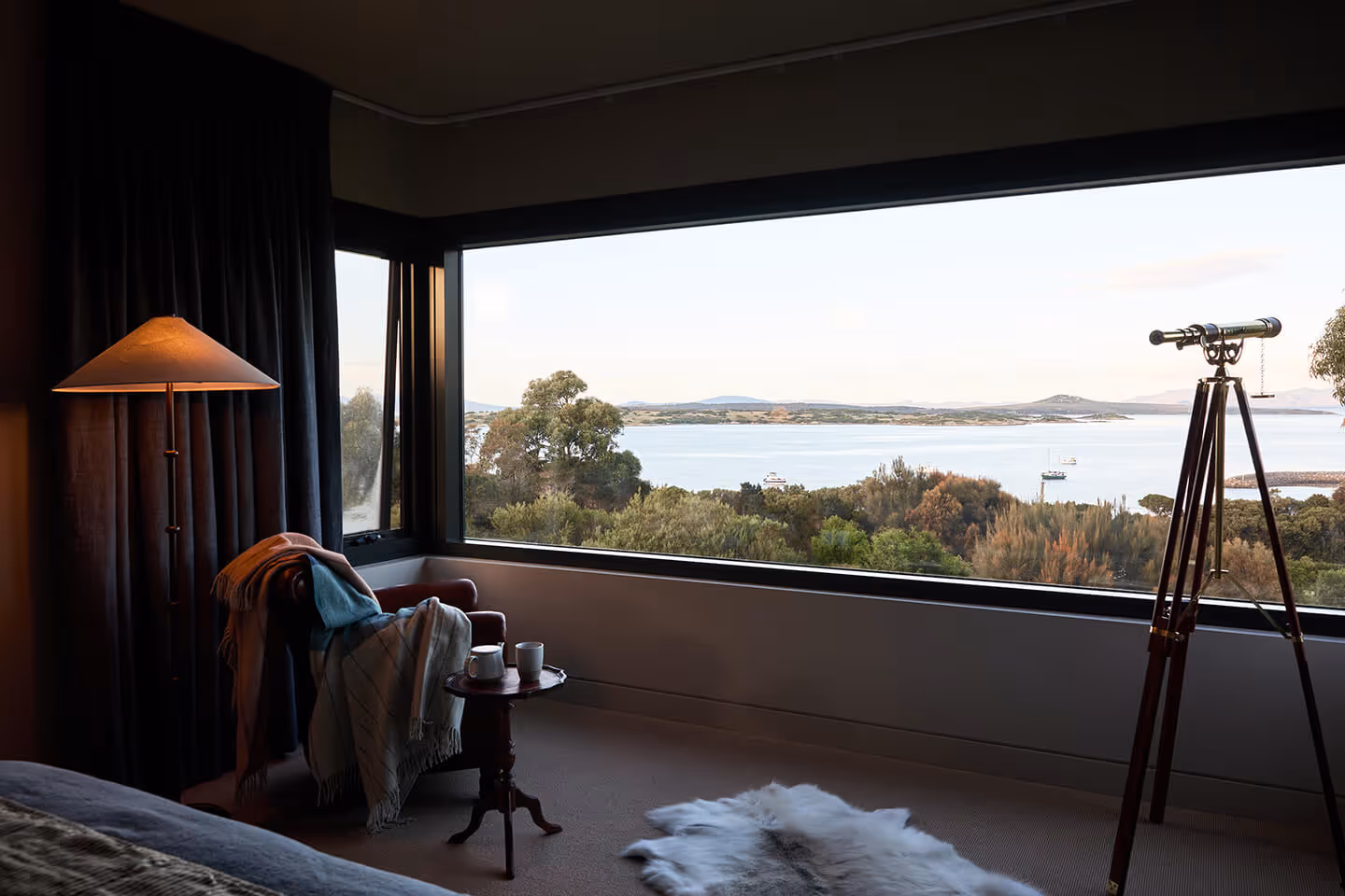 Cozy room with armchair, side table, floor lamp, telescope, and large window showing water and trees.