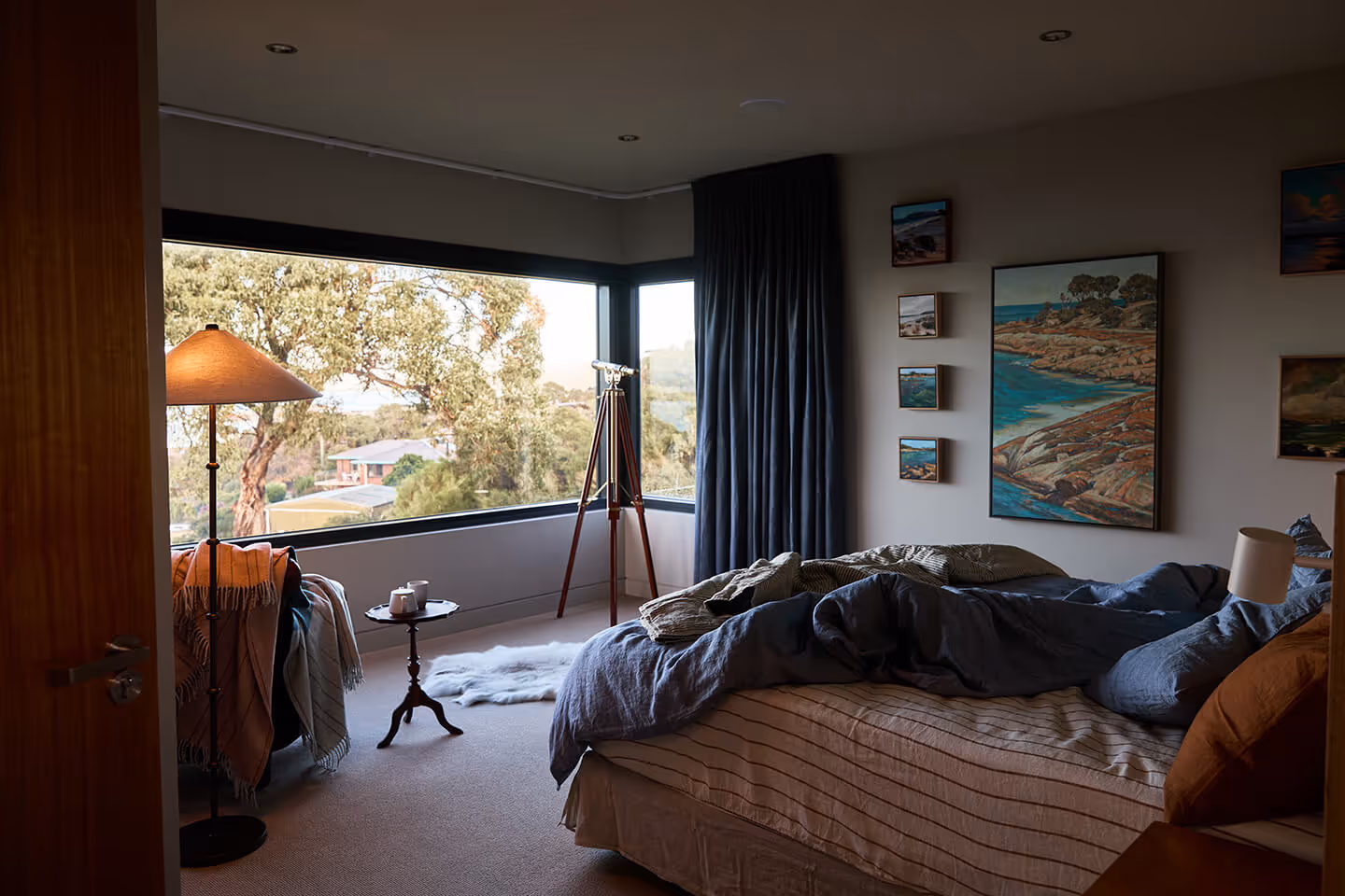 Cozy bedroom with unmade bed, large corner window, floor lamp, telescope, and ocean landscape paintings.