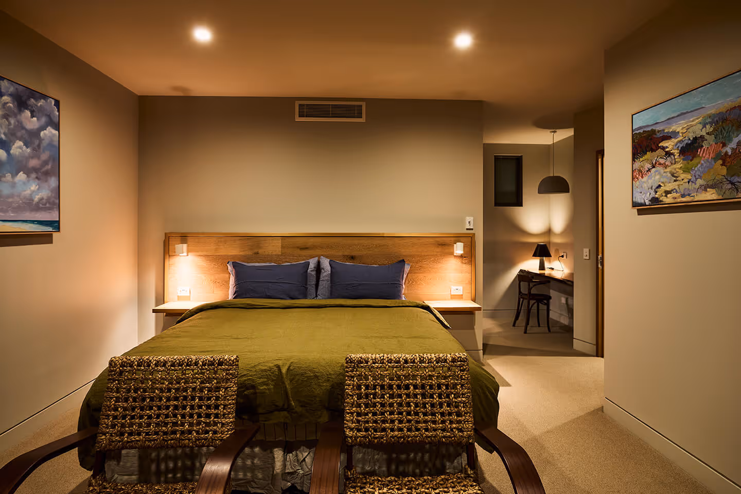 Cozy bedroom with a queen bed, green bedspread, wicker chairs, and framed paintings on beige walls.