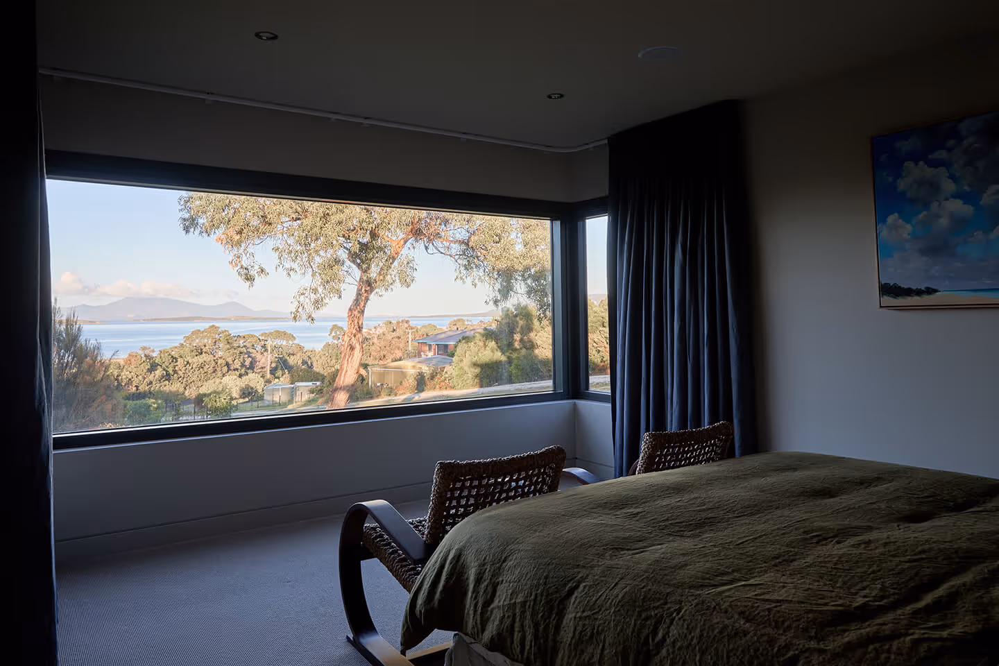 Cozy bedroom with large window overlooking trees, water, and mountains during daytime.