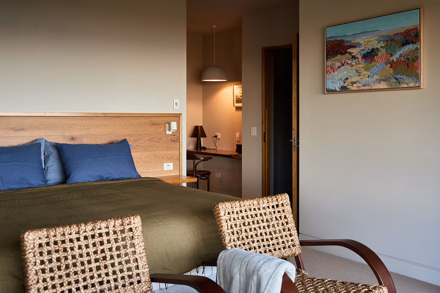 Bedroom with green bedspread, blue pillows, woven chairs, desk with lamps, and landscape painting.